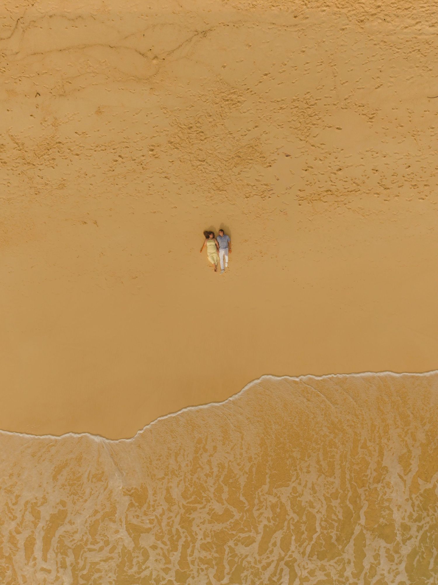 Aerial view of two people walking on a vast golden sandy beach near the shoreline.