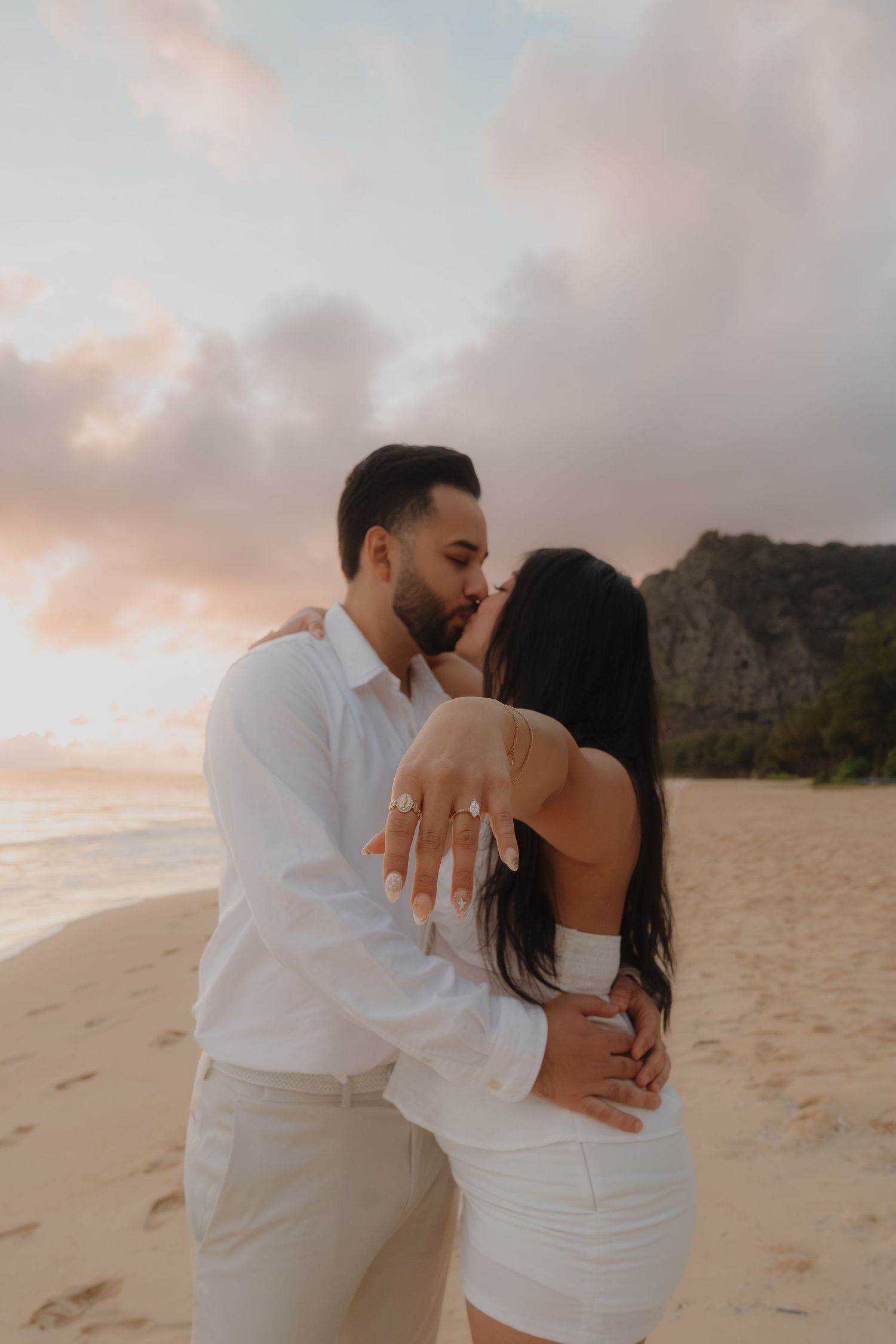 Engaged couple kisses on a beach at sunset, woman showing off her diamond engagement ring.