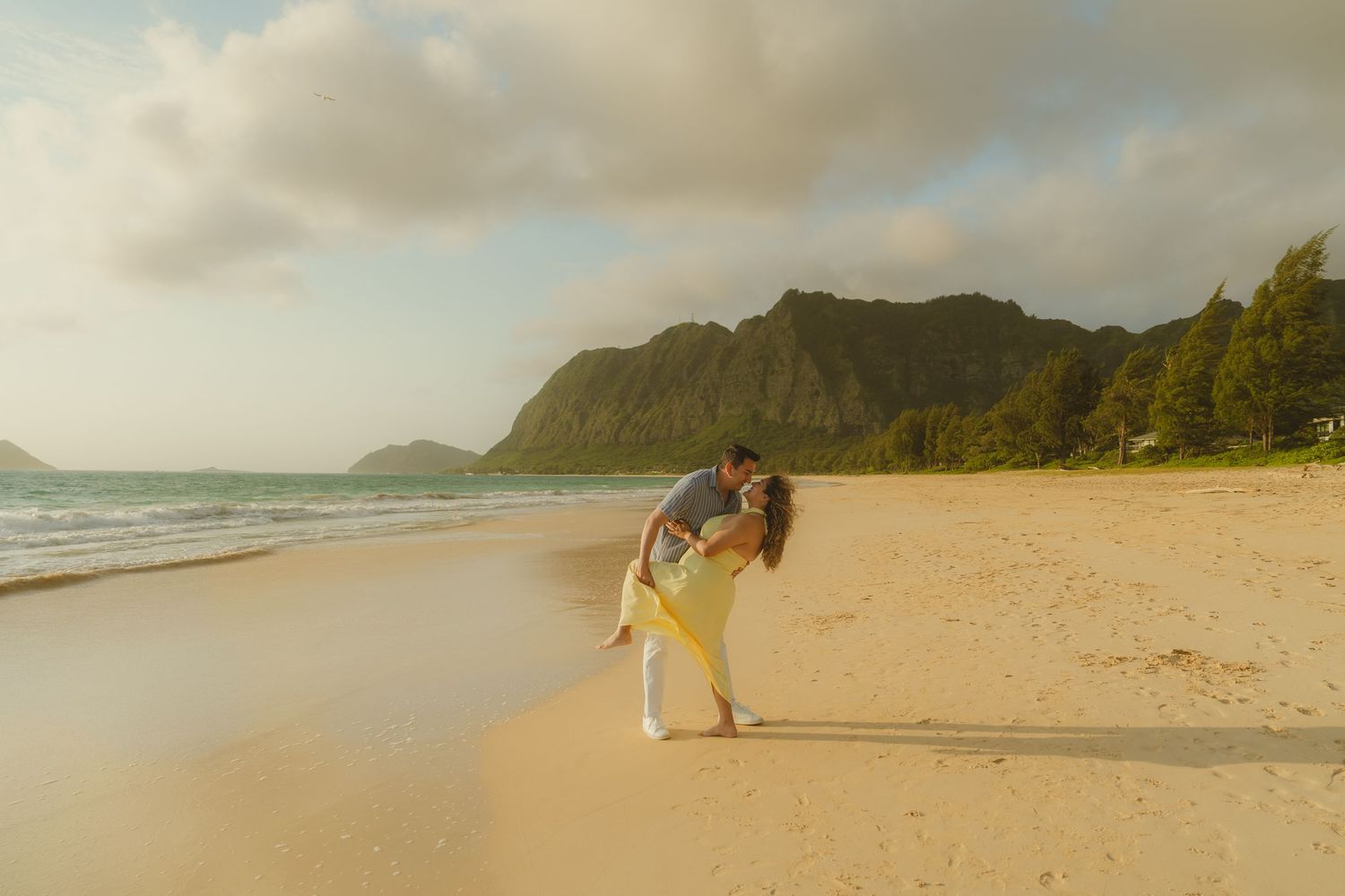 Couple sharing a romantic dip kiss on a sandy Hawaiian beach at golden hour with lush green mountains in the background.