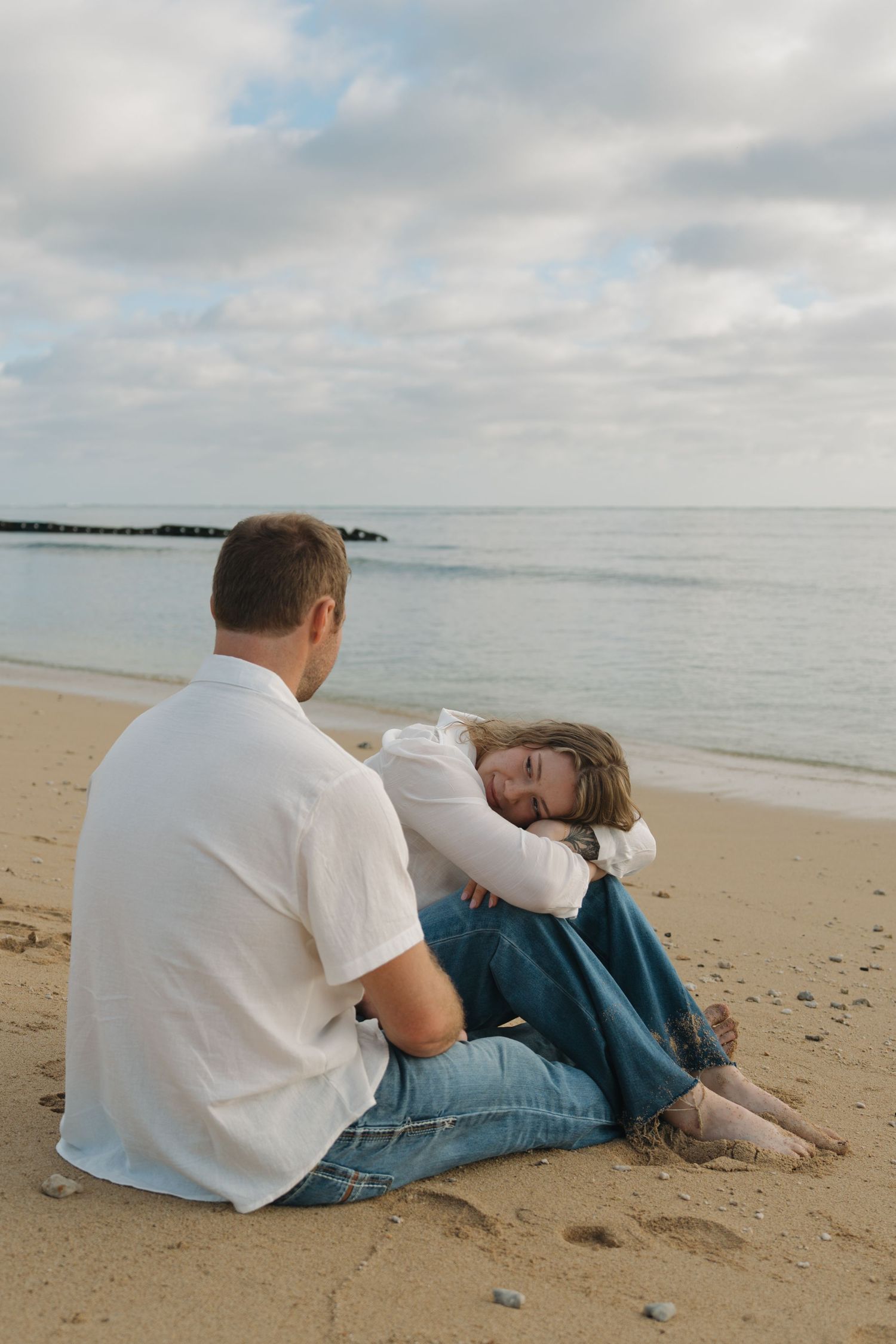 Couple sitting on sandy beach at dusk, woman resting her head on mans shoulder.