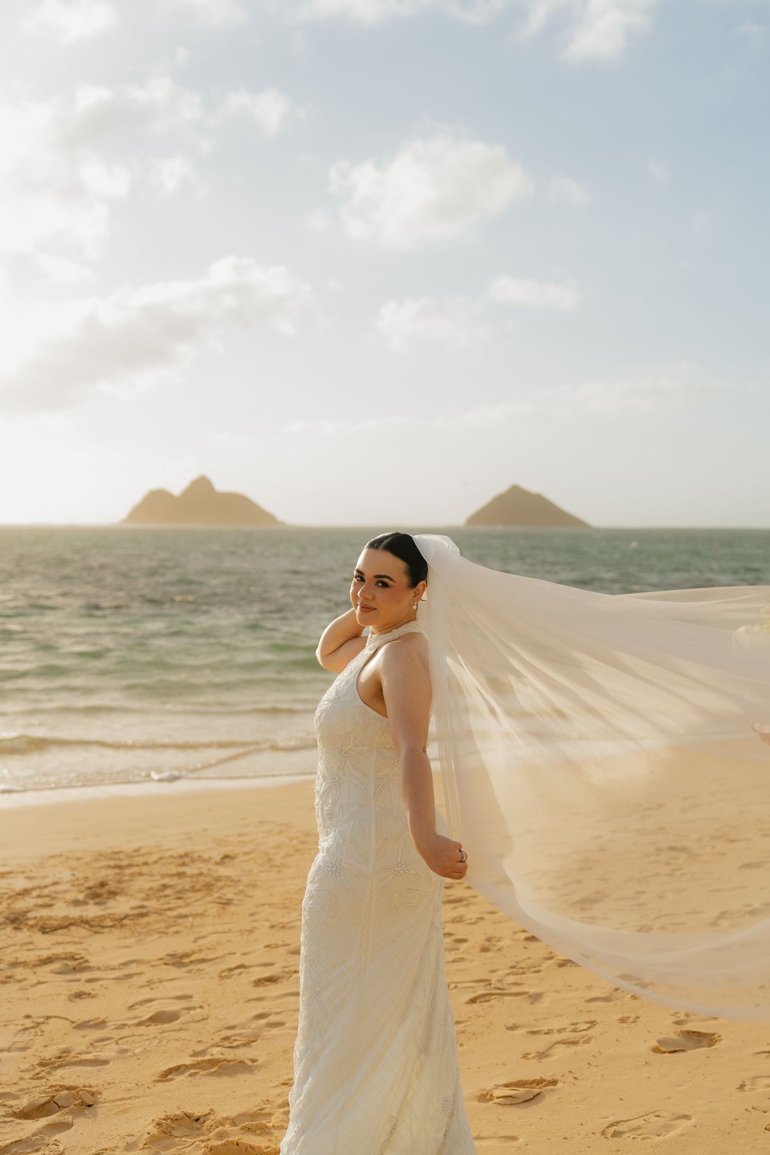 Bride in white lace gown with flowing veil on sandy beach, Mokulua Islands visible in ocean background, Hawaii.