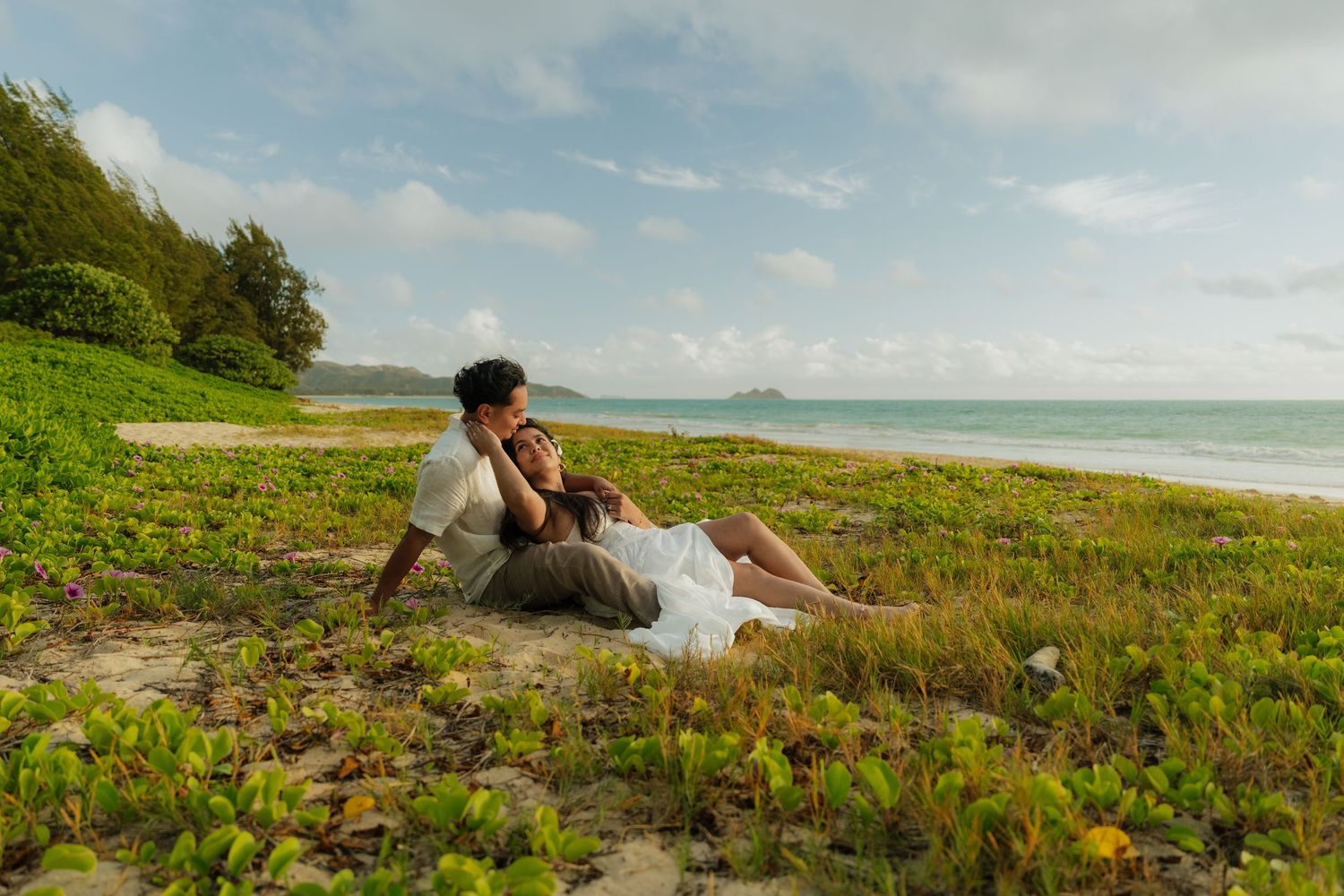 Couple relaxing on a lush green tropical beach shore, leaning together romantically with ocean waves in the background.