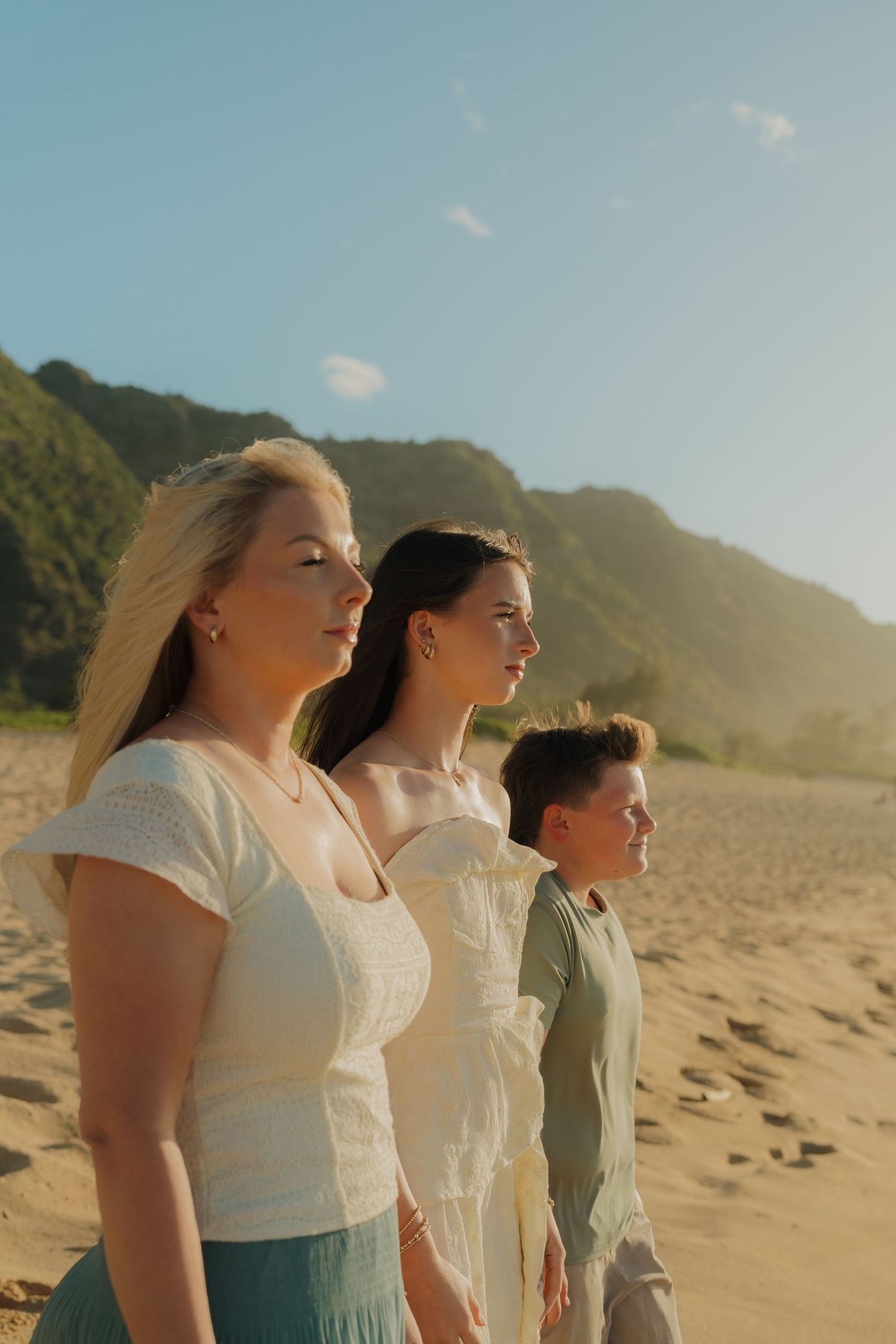 Two women and a boy standing on sandy beach with green mountains at sunset.