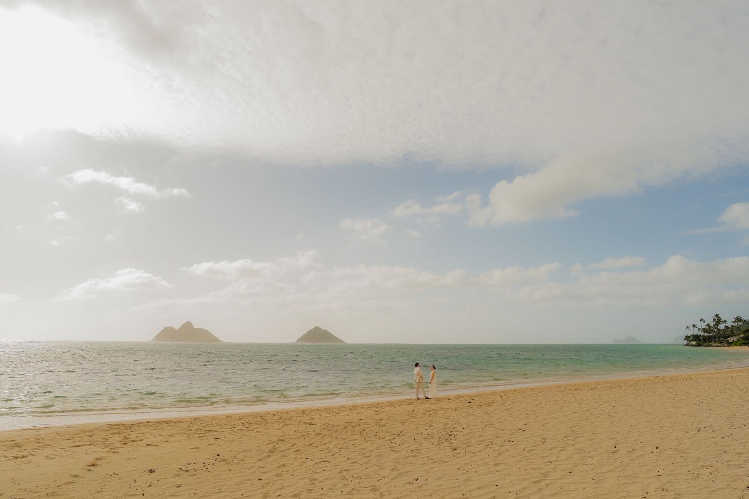 Couple walking on Lanikai Beach in Hawaii with Mokulua Islands in the background under a bright sky.