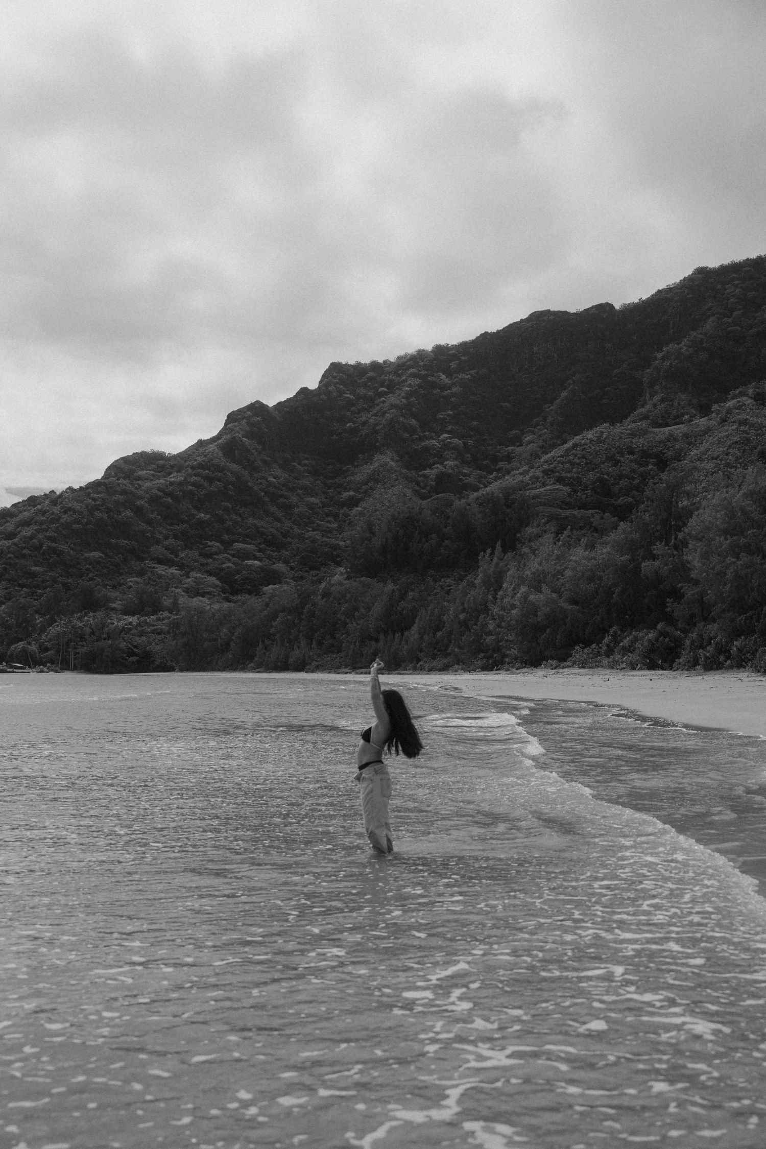 Black and white photo of woman standing in ocean waves with lush mountain landscape behind her.