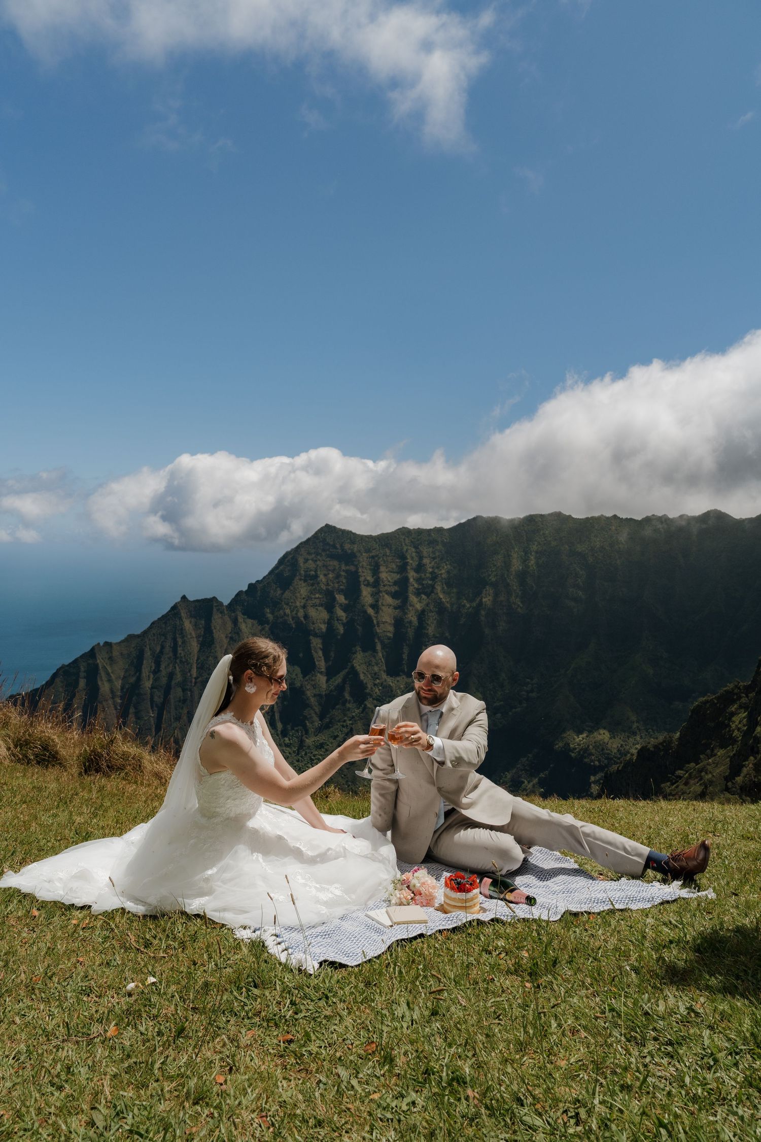 Bride and groom share a champagne toast during an elopement picnic on a lush hillside with stunning mountain views on Kauai Hawaii