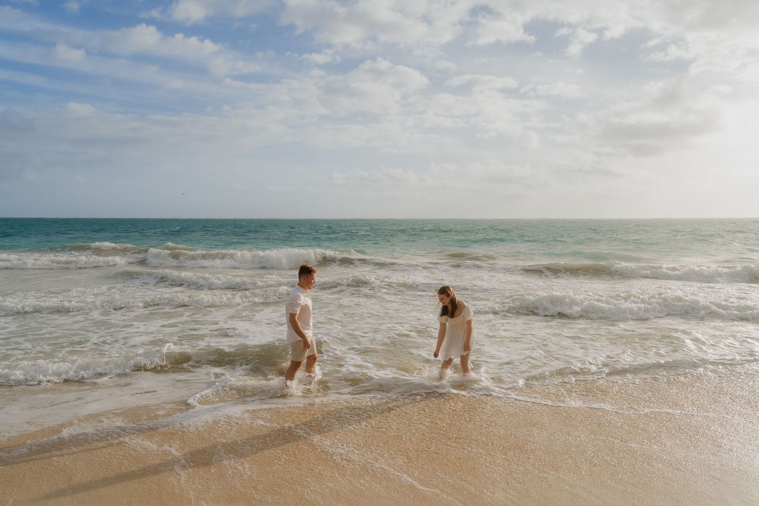 Couple wading through ocean waves on a sandy beach under a partly cloudy sky.