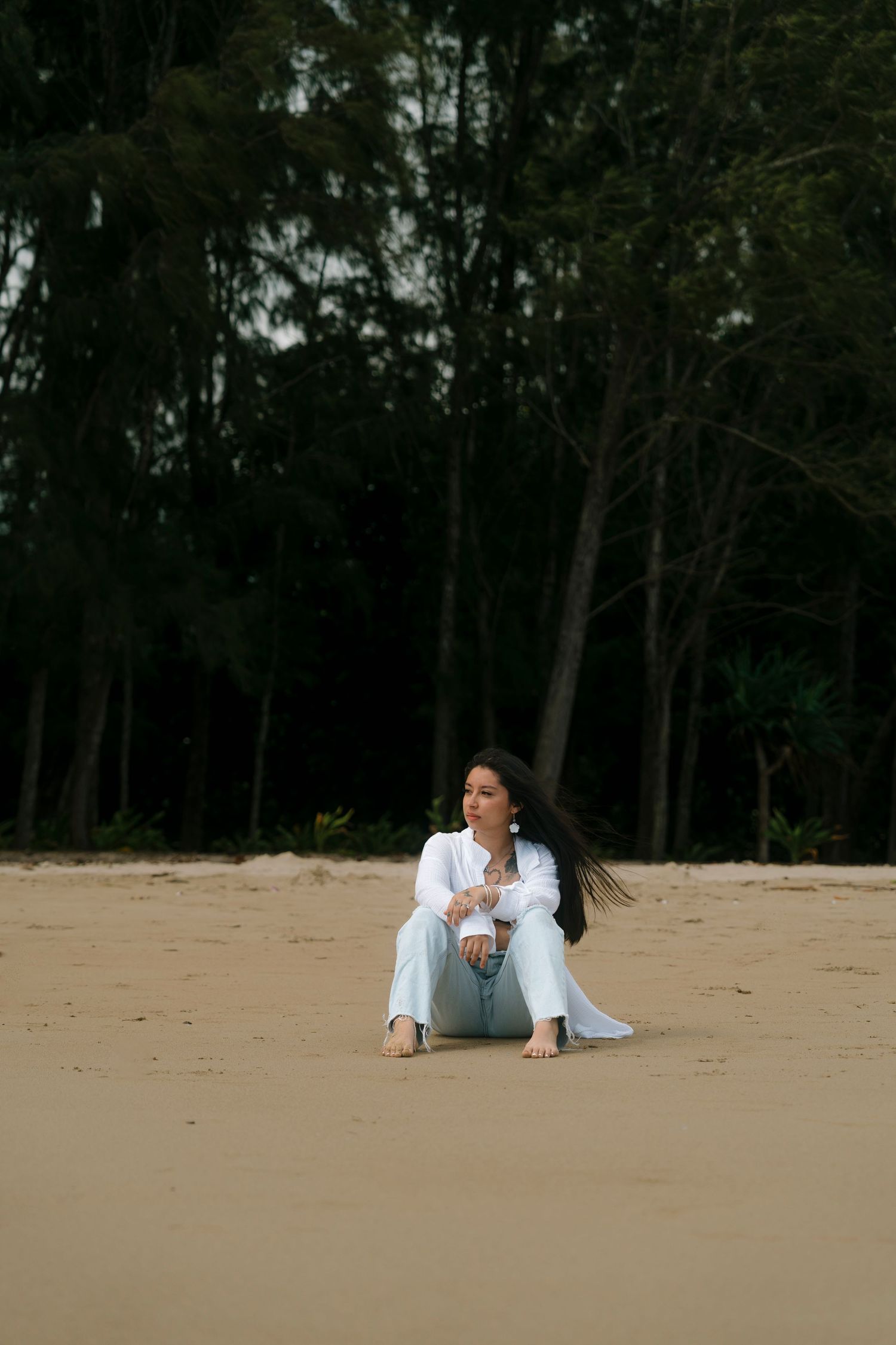 Woman in white shirt and jeans sitting on sandy beach with dark forest background.
