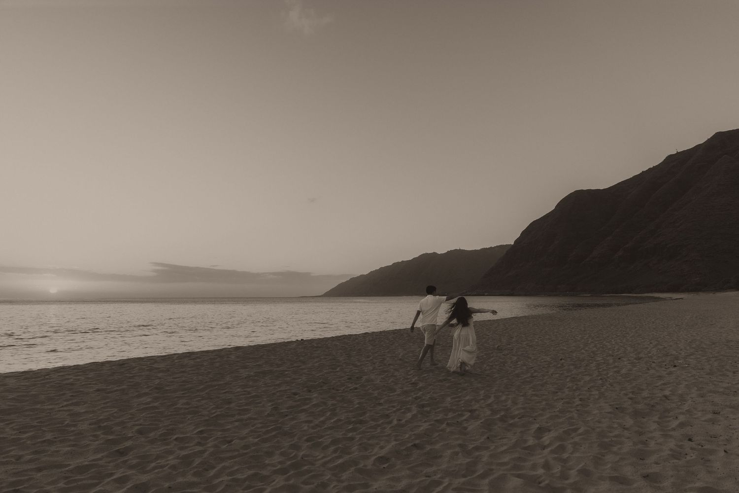 Couple walking hand in hand on a sandy beach at sunset with mountains in the background, sepia tone.