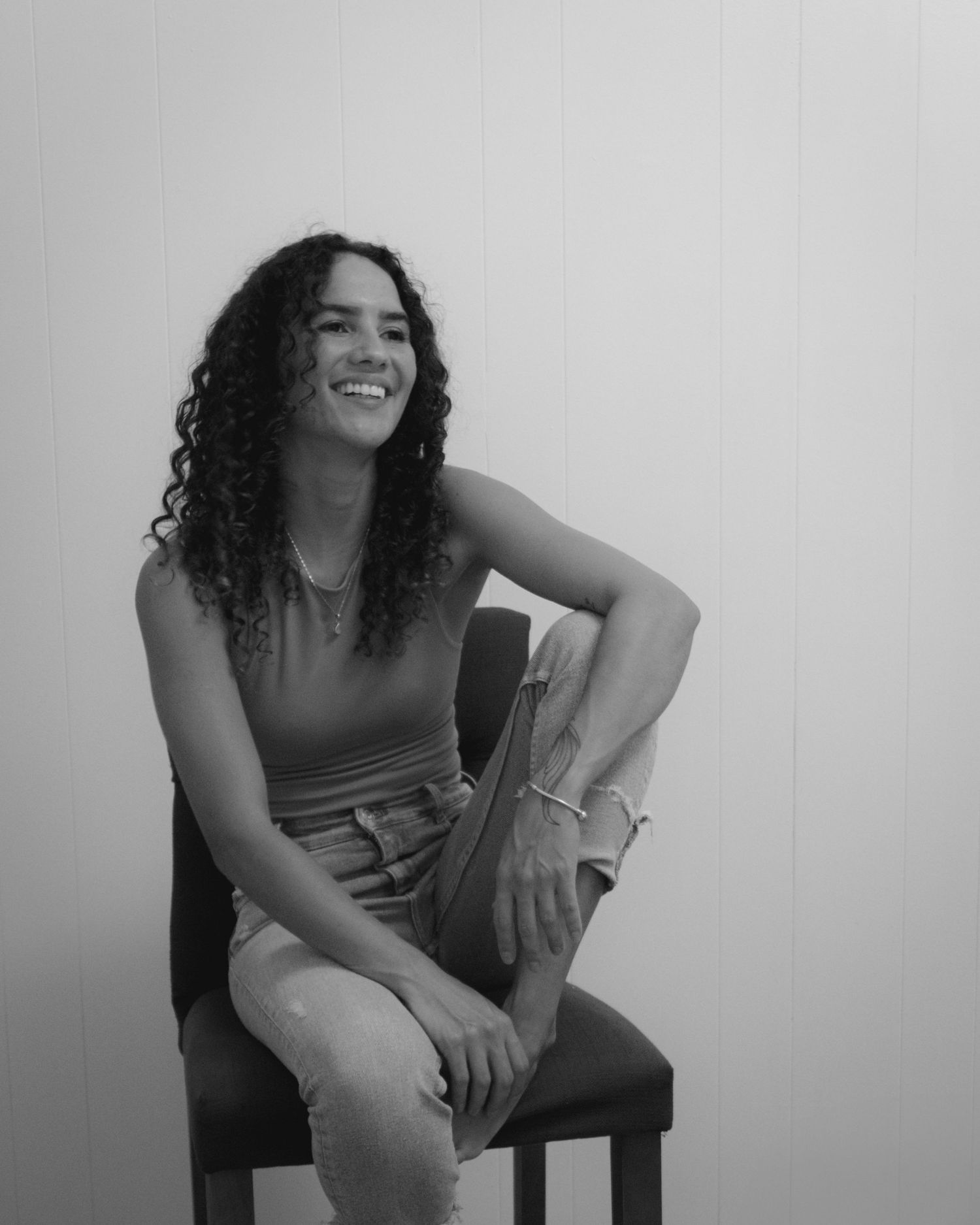 Black and white portrait of a smiling young woman with curly hair sitting on a chair.