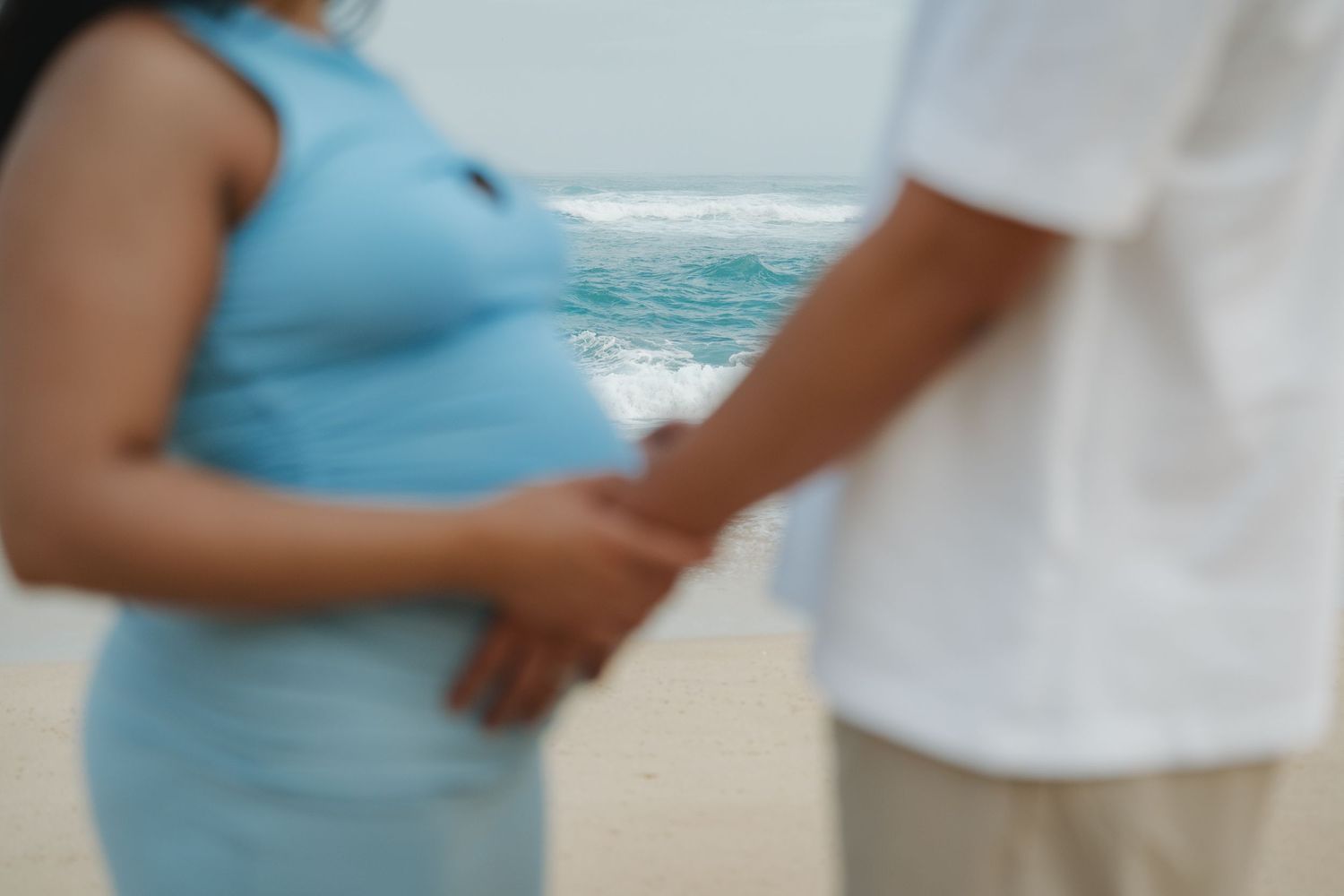 Close-up of pregnant woman in blue dress holding partner's hand on beach.