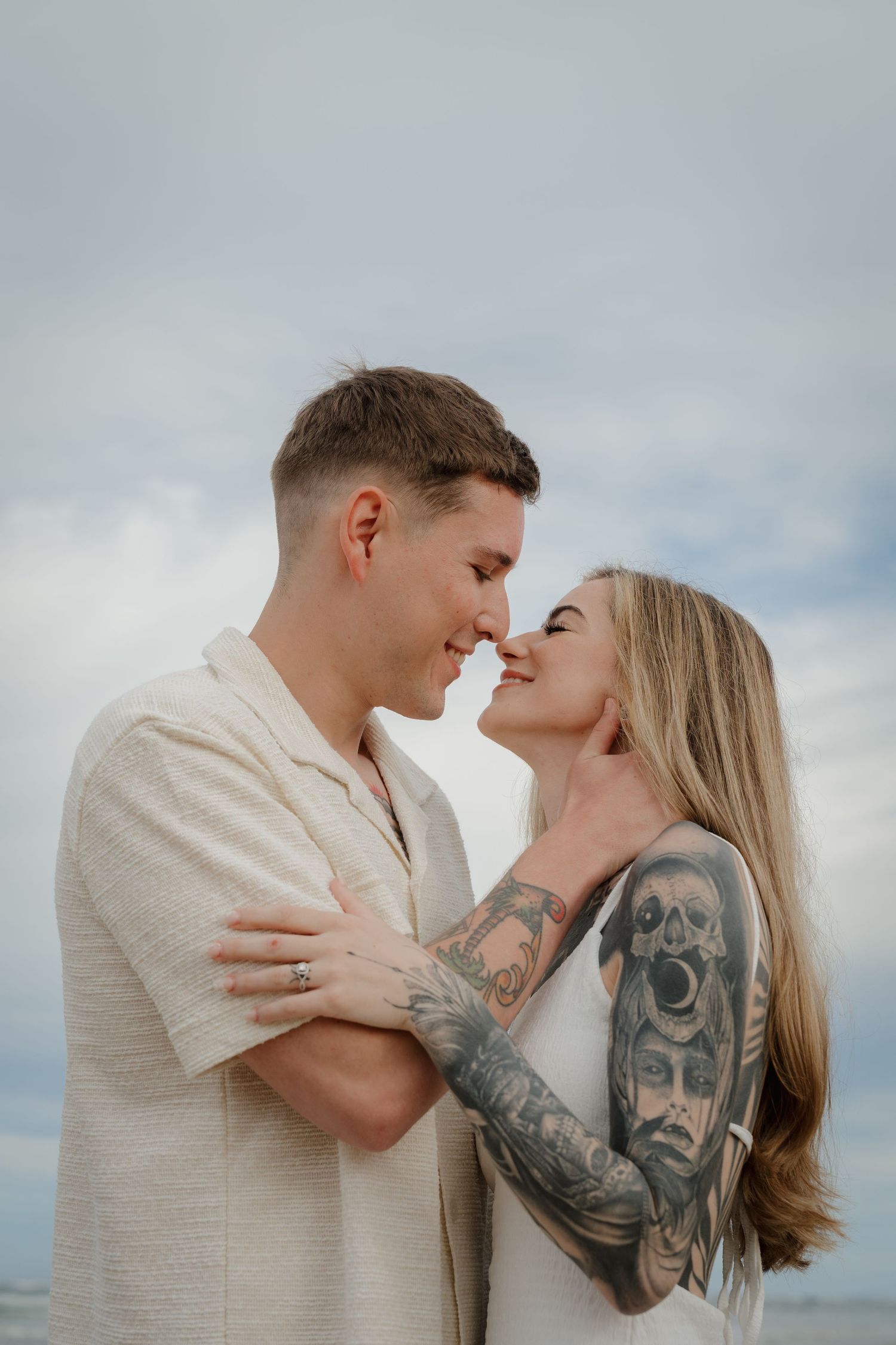 Tattooed couple sharing intimate nose-to-nose moment embracing under cloudy sky at beach.