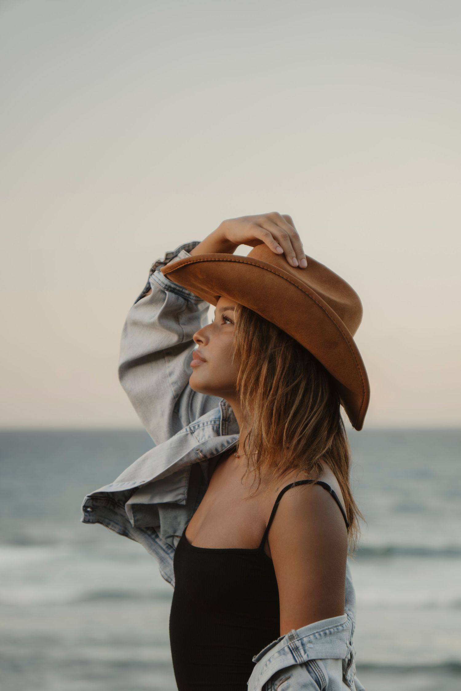 Young woman in brown cowboy hat and denim jacket posing at beach during sunset.