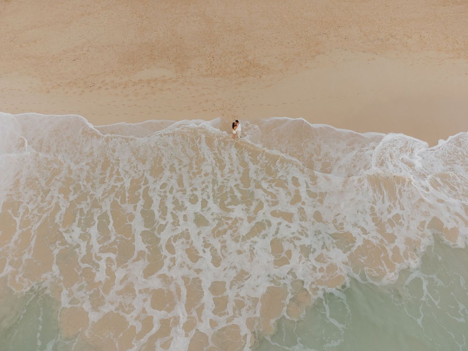Aerial view of a couple standing at the ocean's edge on a sandy beach with white foam waves.