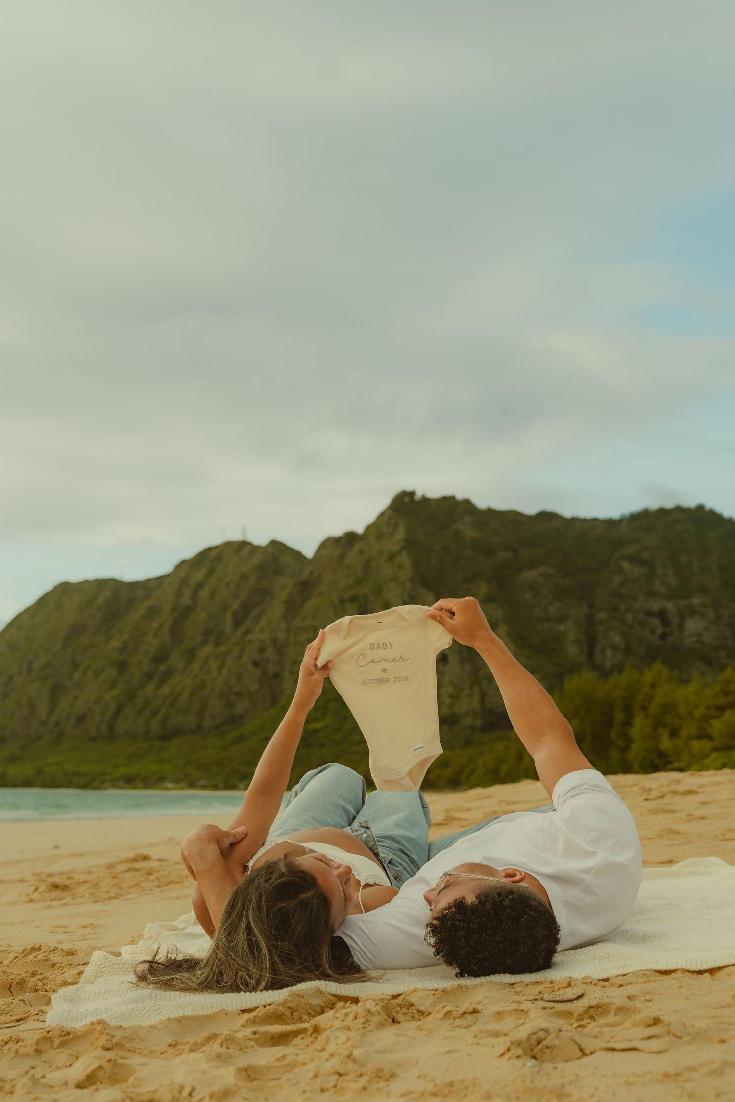 Couple lying on sandy beach holding up a baby onesie, with lush green mountains and ocean in the background.