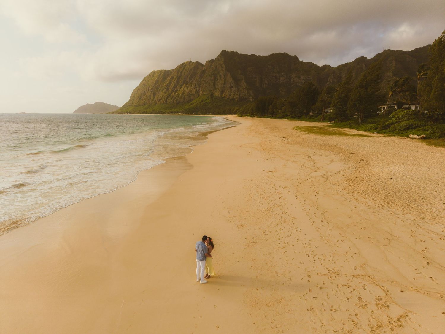 Aerial view of a couple walking on a golden sandy beach in Hawaii with dramatic green mountains and ocean waves.