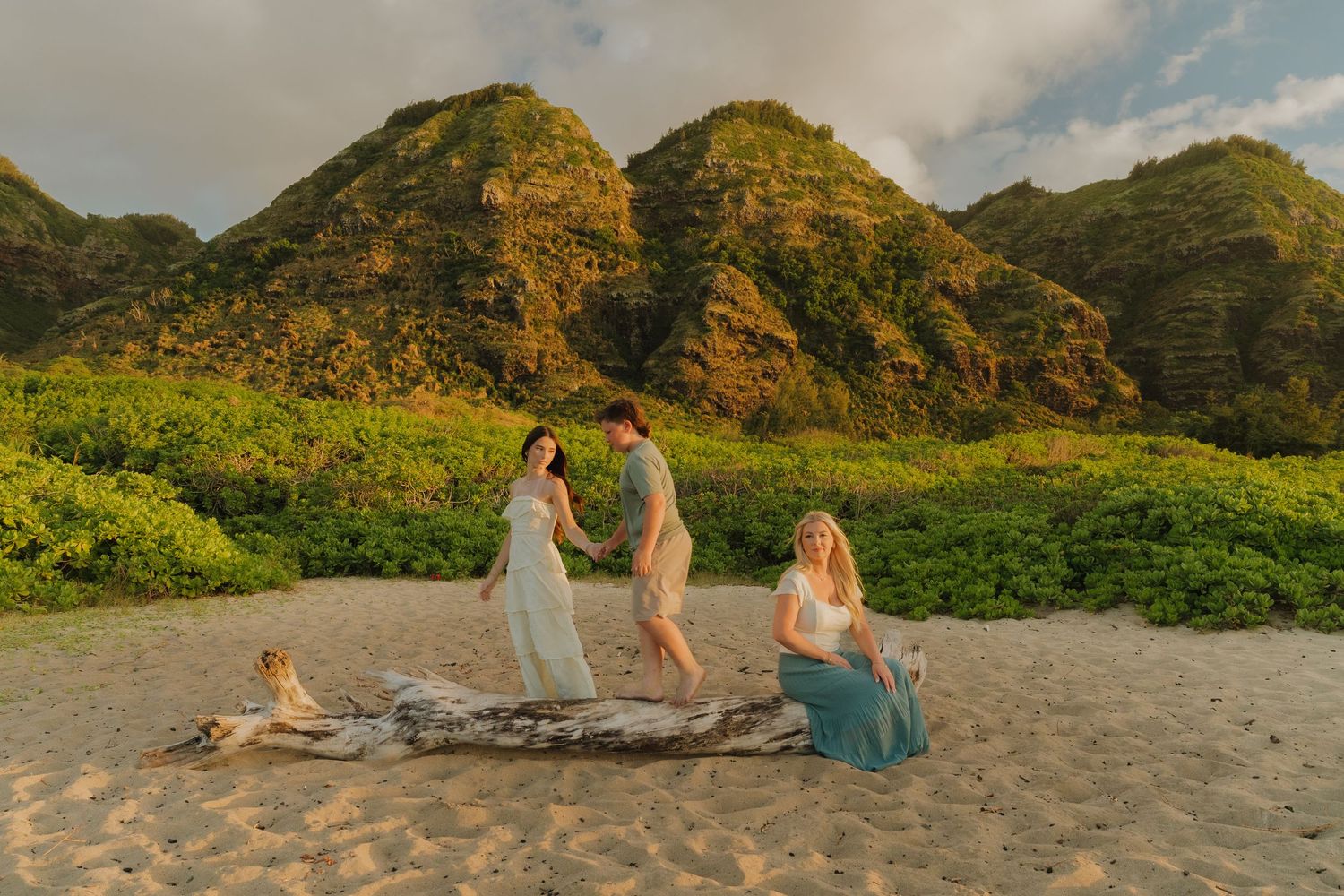 Couple holds hands on driftwood at golden hour on a Hawaiian beach with lush green mountains backdrop.