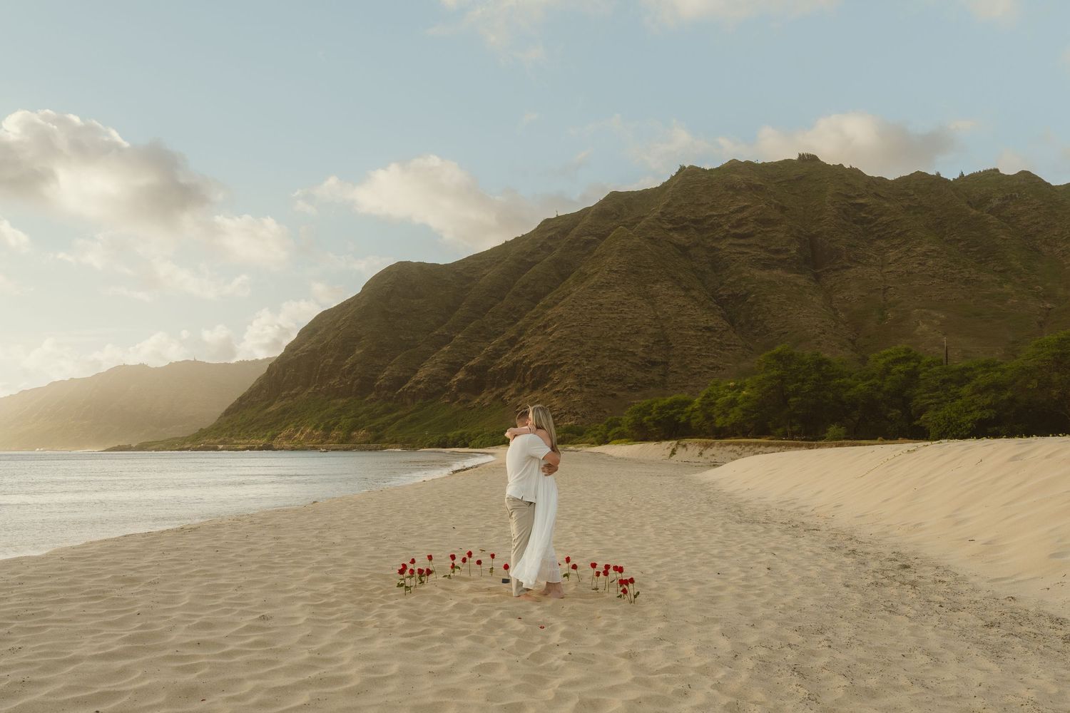 Couple embracing on a sandy Hawaiian beach at sunset with red flowers and lush green mountains in the background.