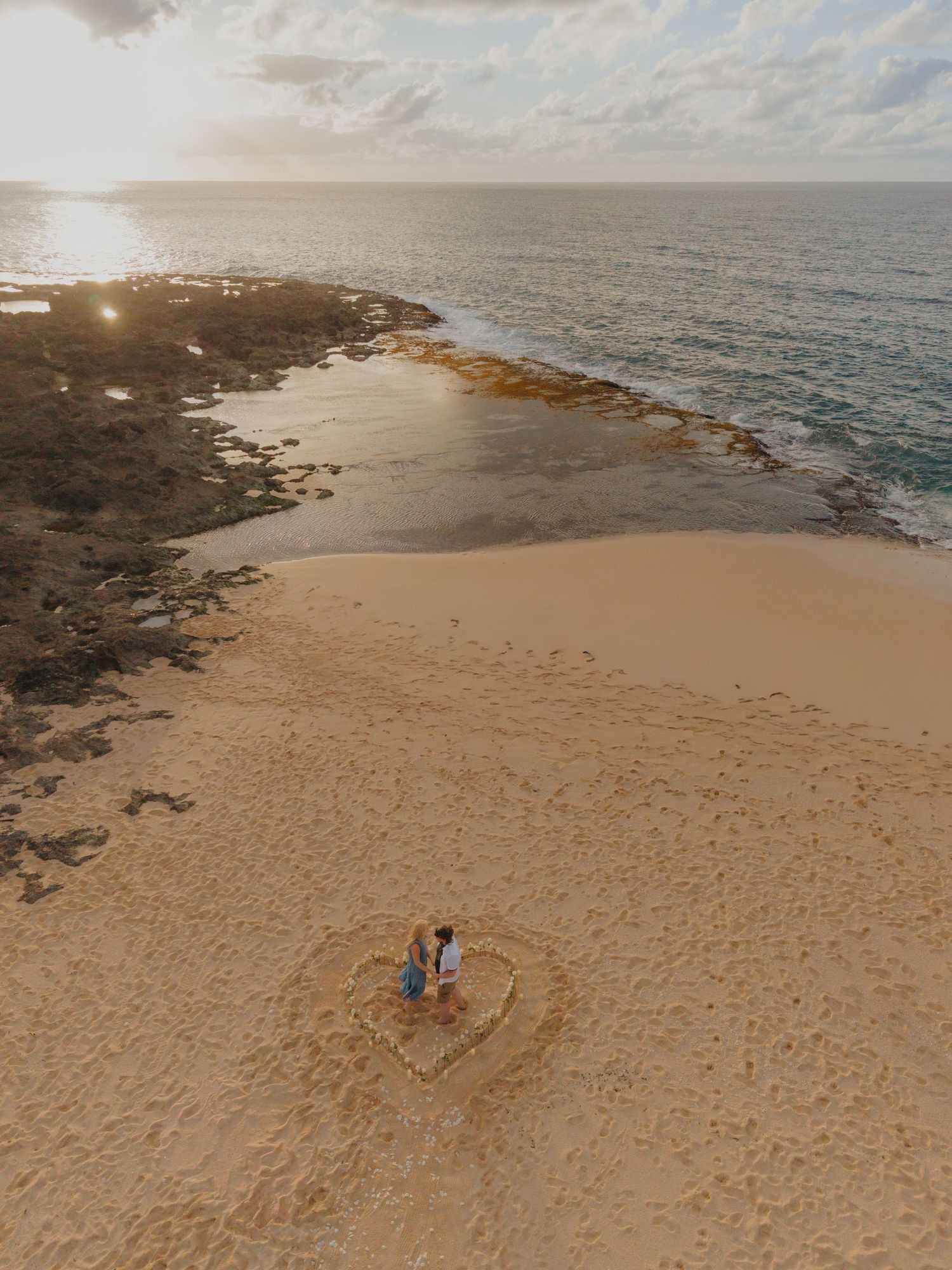 Aerial drone view of a couple standing inside a heart drawn in golden sand on a beach at sunset near rocky shores. Hawaii proposal roses