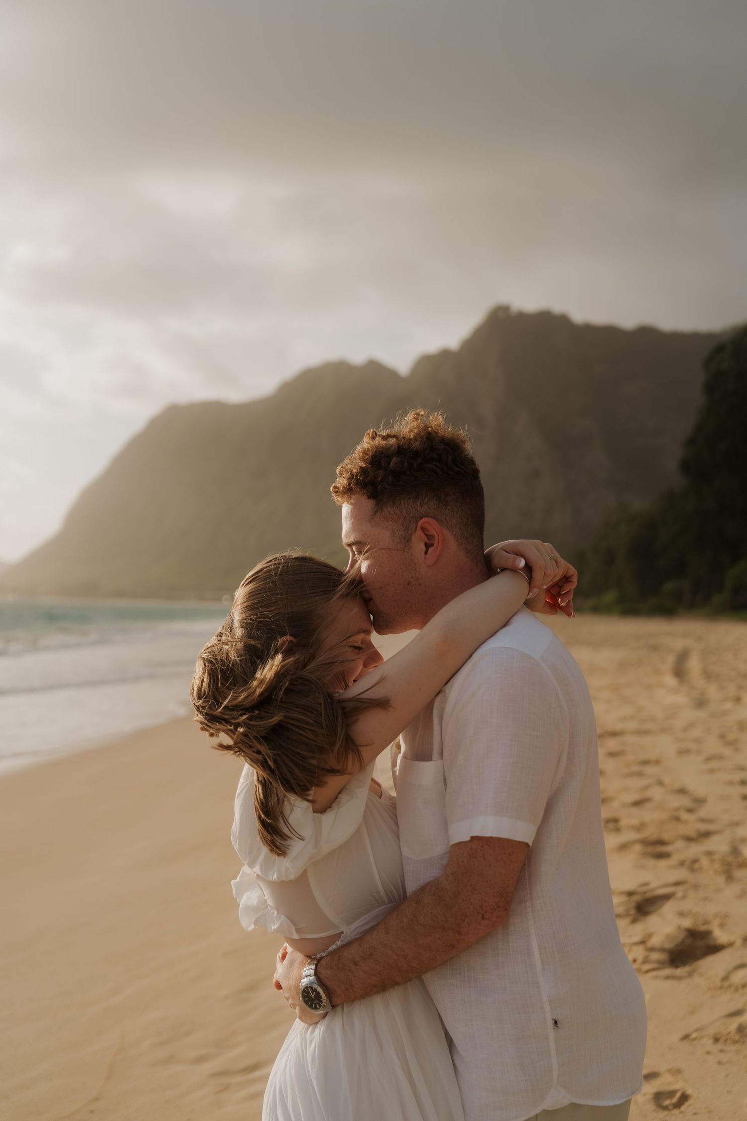 Couple sharing a romantic kiss on a beach with dramatic mountain backdrop at golden sunset.
