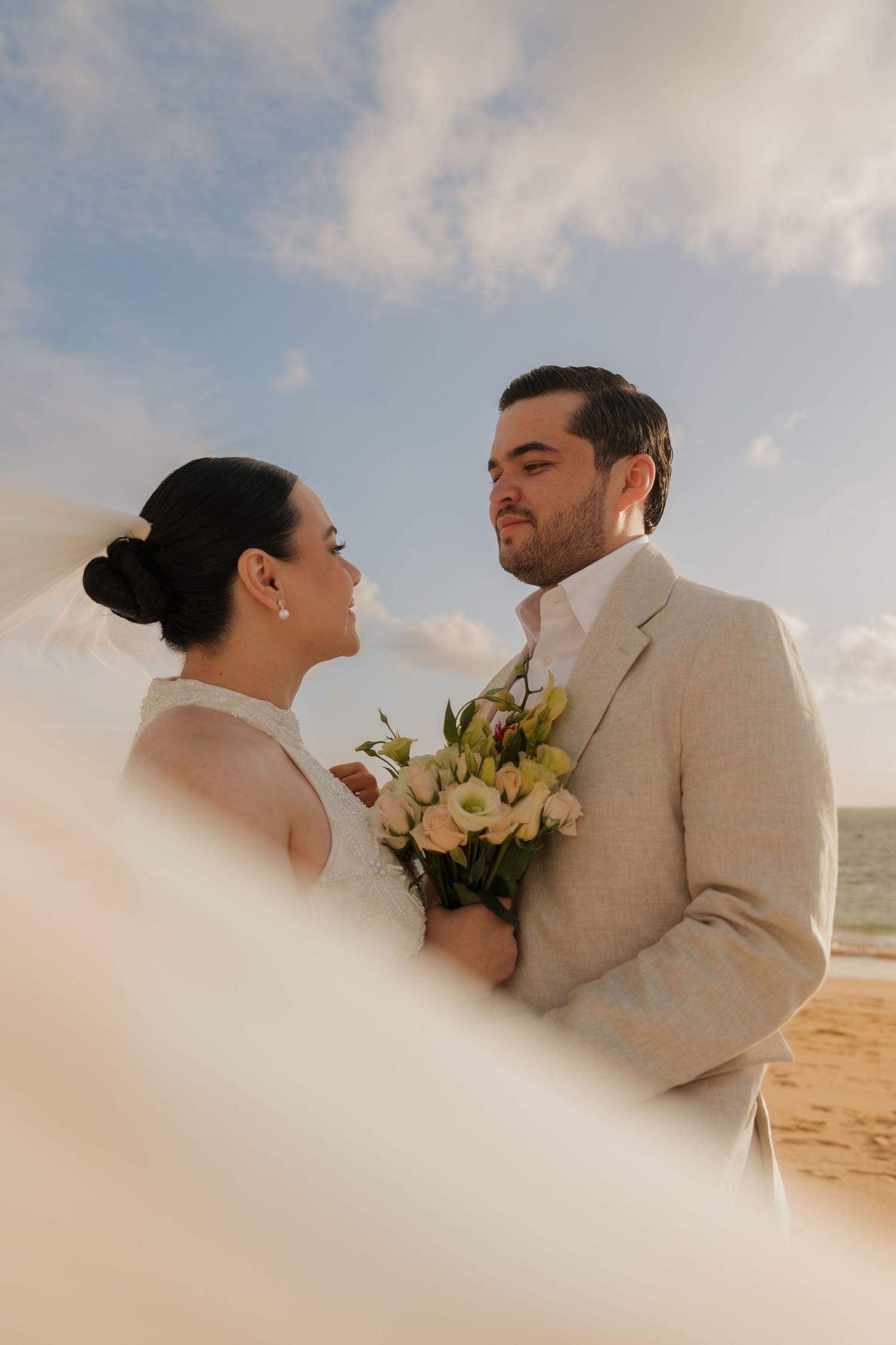 Bride and groom share a romantic moment on a beach, holding a white bouquet with the ocean and blue sky behind them.