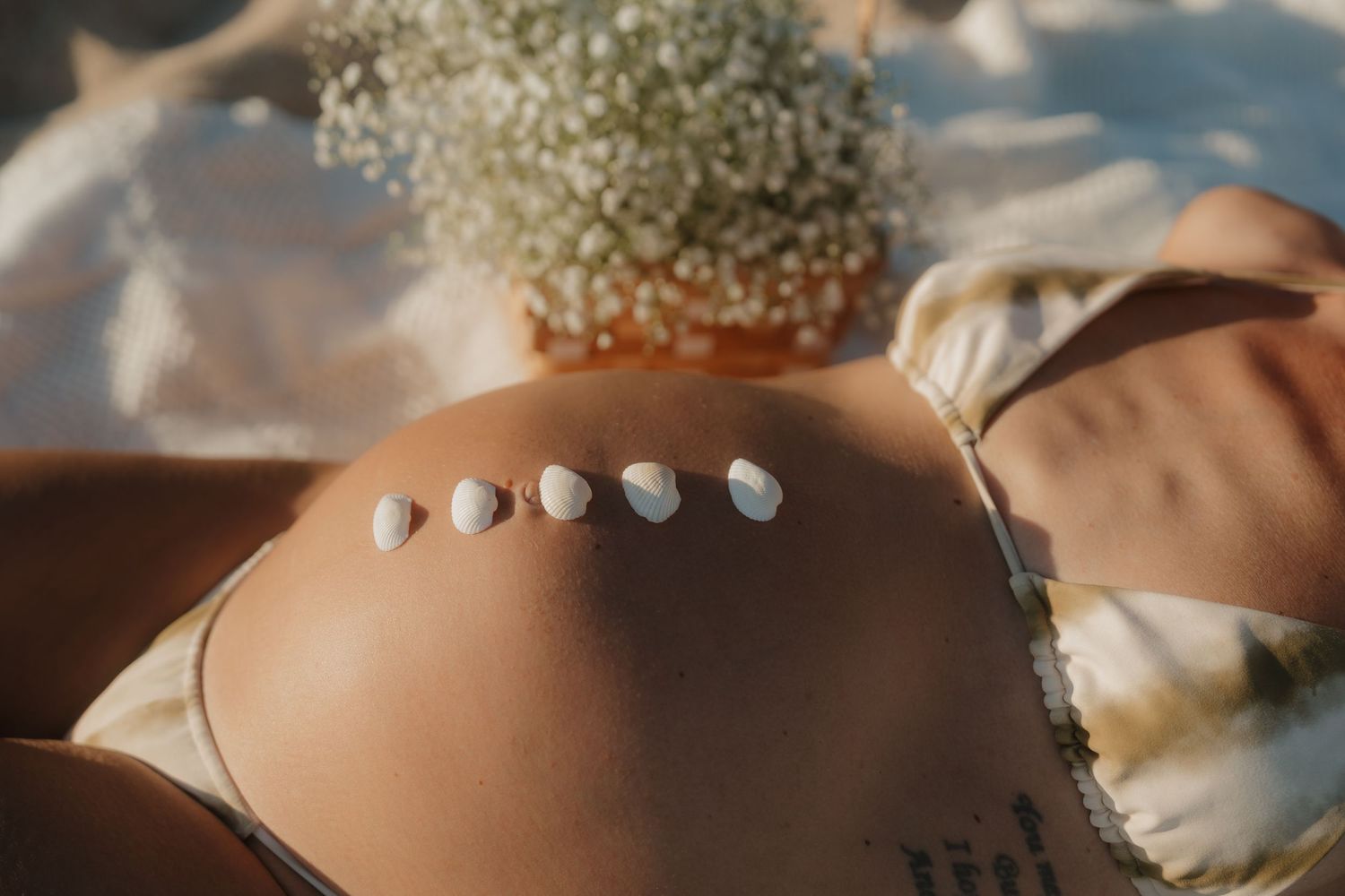 Row of small seashells arranged on pregnant belly with baby's breath flowers in background.