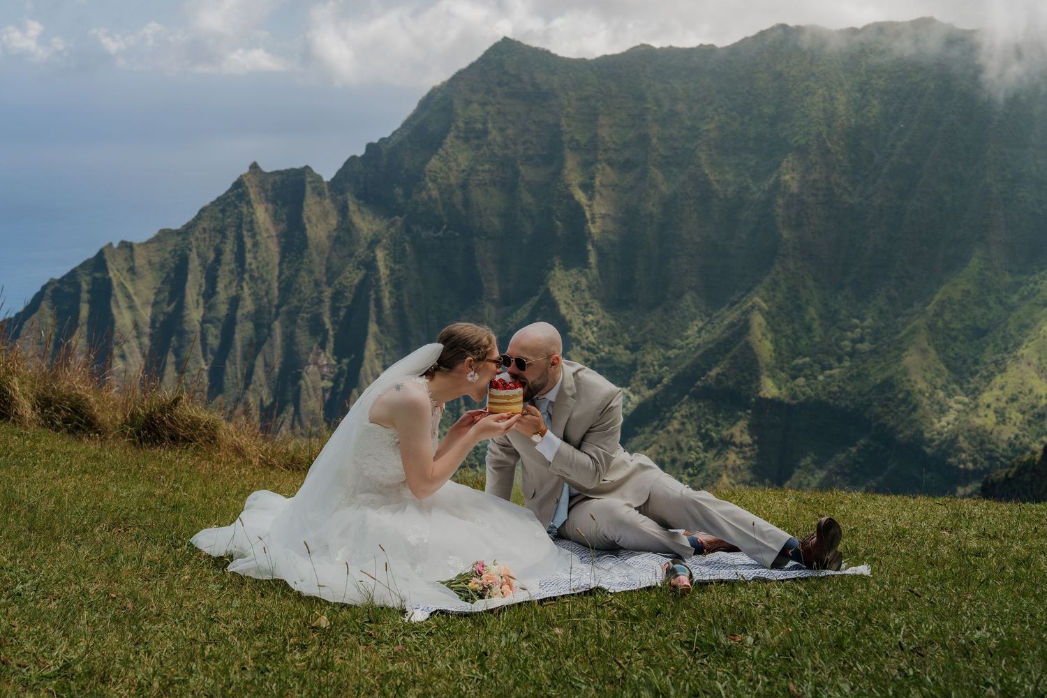 Bride and groom toasting champagne on a scenic Hawaiian cliffside with dramatic Na Pali Coast mountains backdrop.