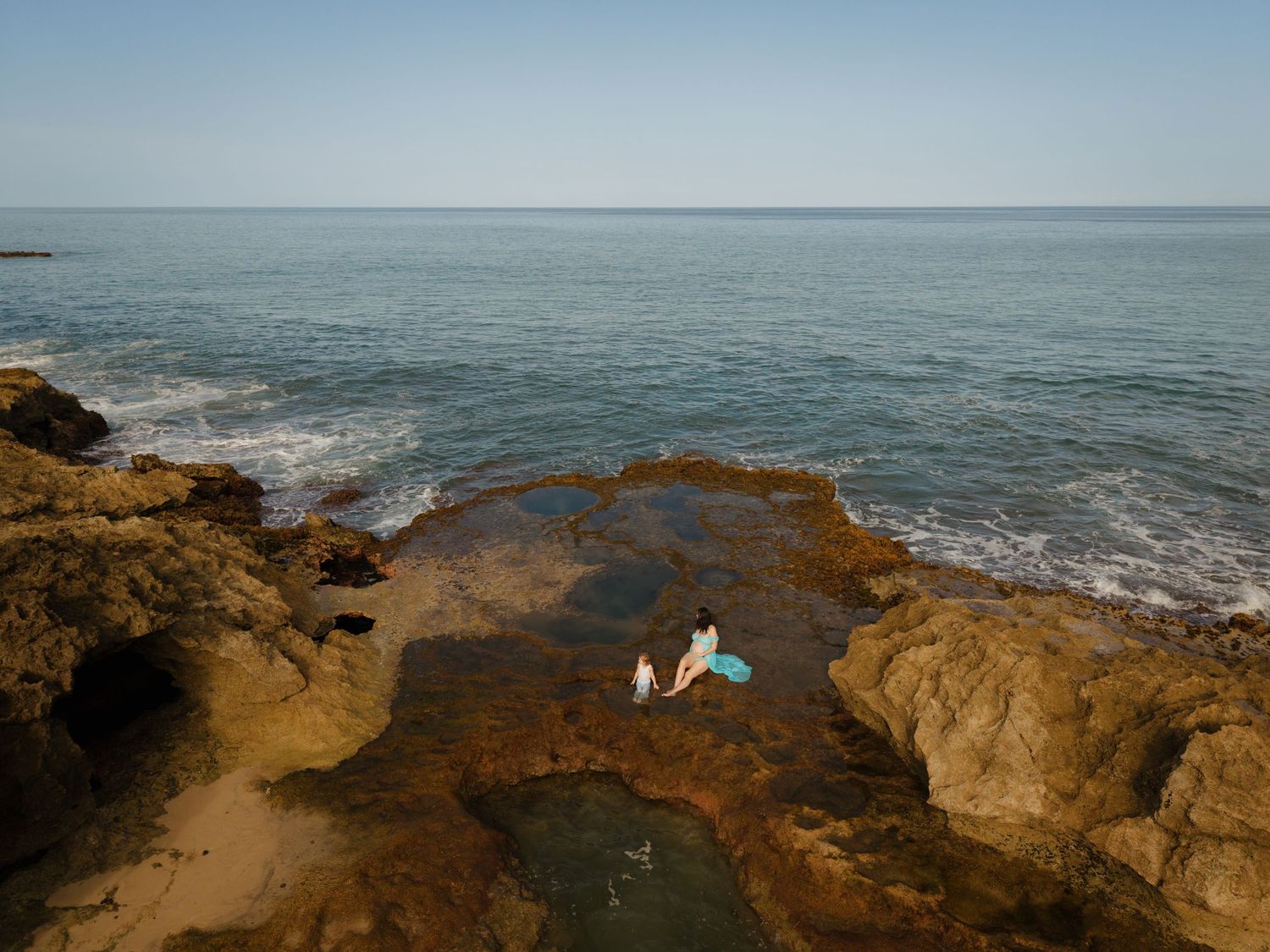 Two people relax in a rocky tidal pool by the ocean on a clear sunny day.
