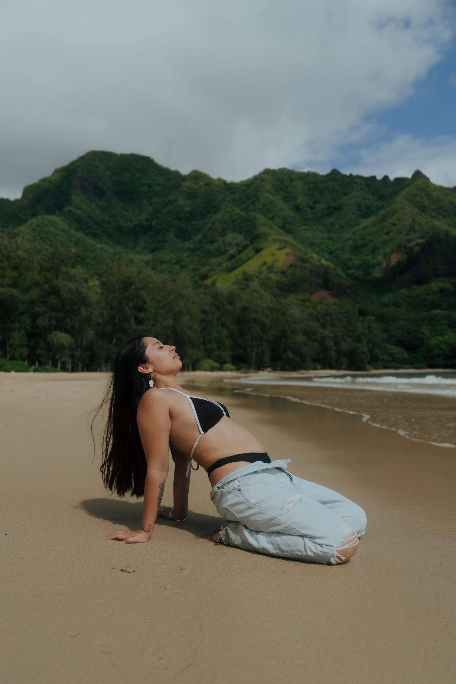 Woman in black crop top and jeans kneeling on sandy beach with lush green mountains behind her.