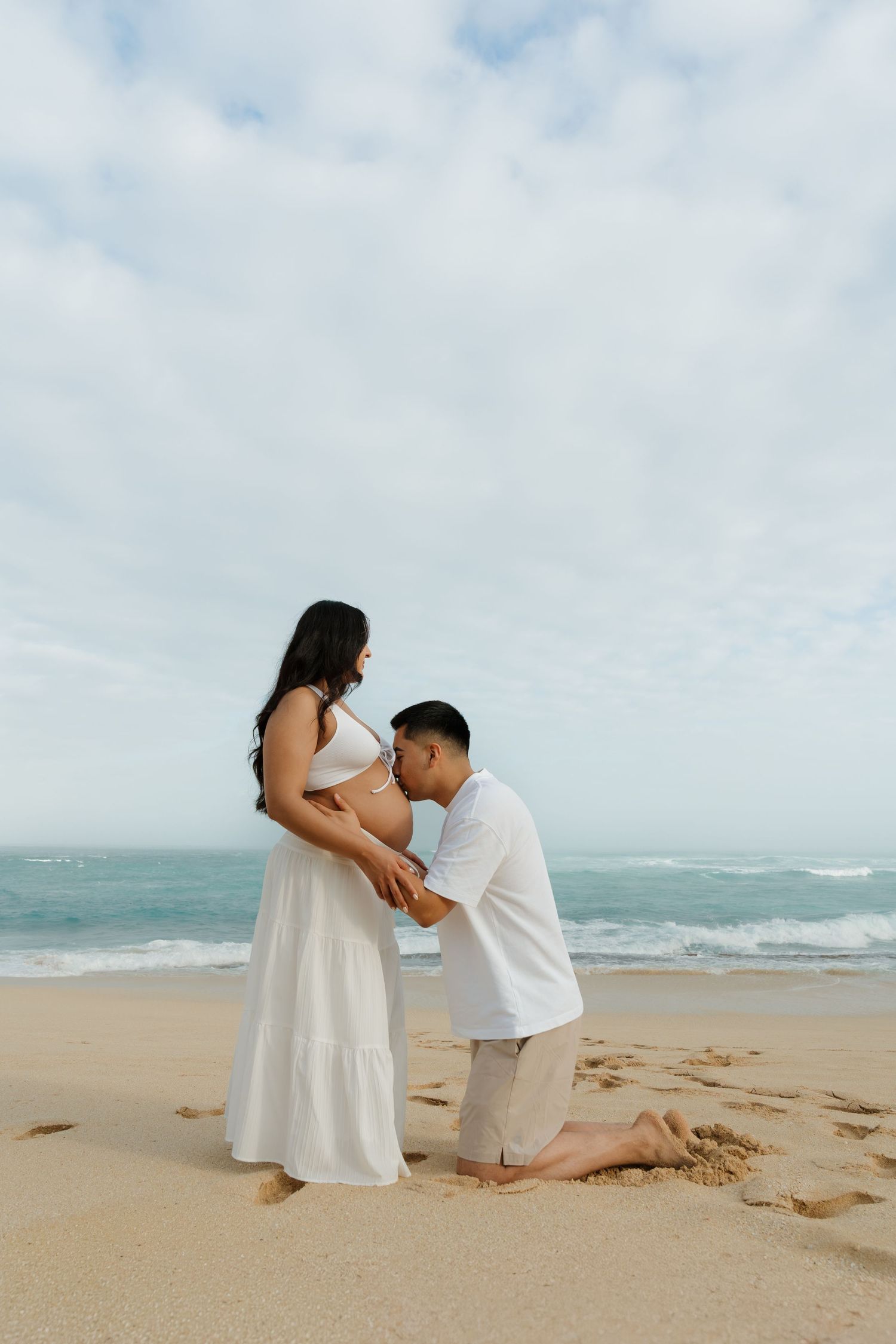 Partner kneeling on sand kissing pregnant woman's belly at the beach.
