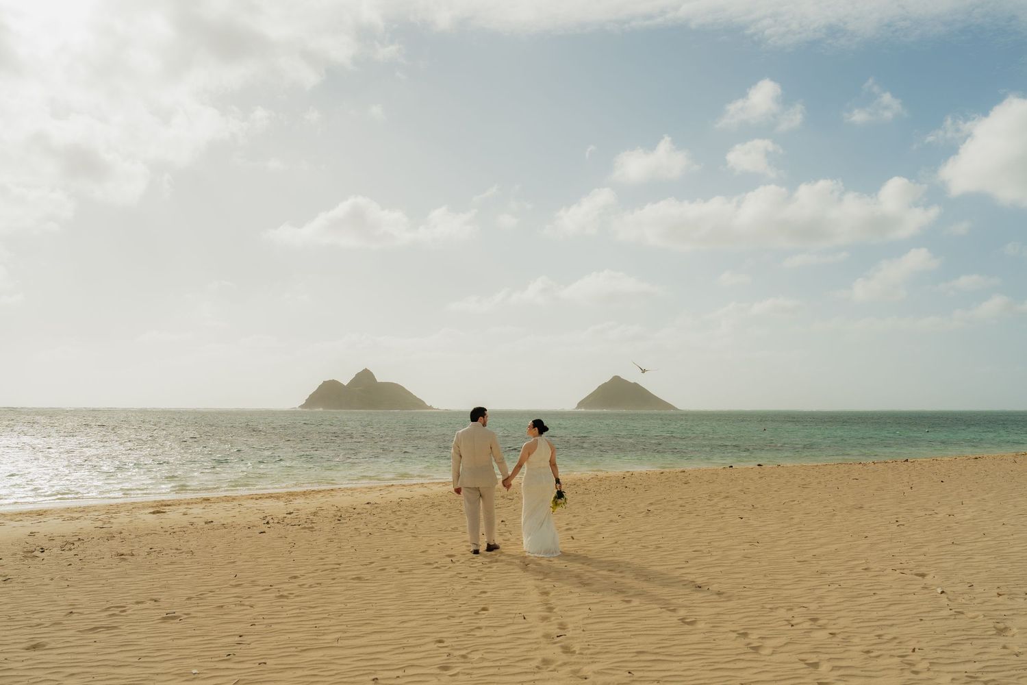 Couple holding hands on a sandy beach in Hawaii with turquoise ocean and twin island peaks in the background.