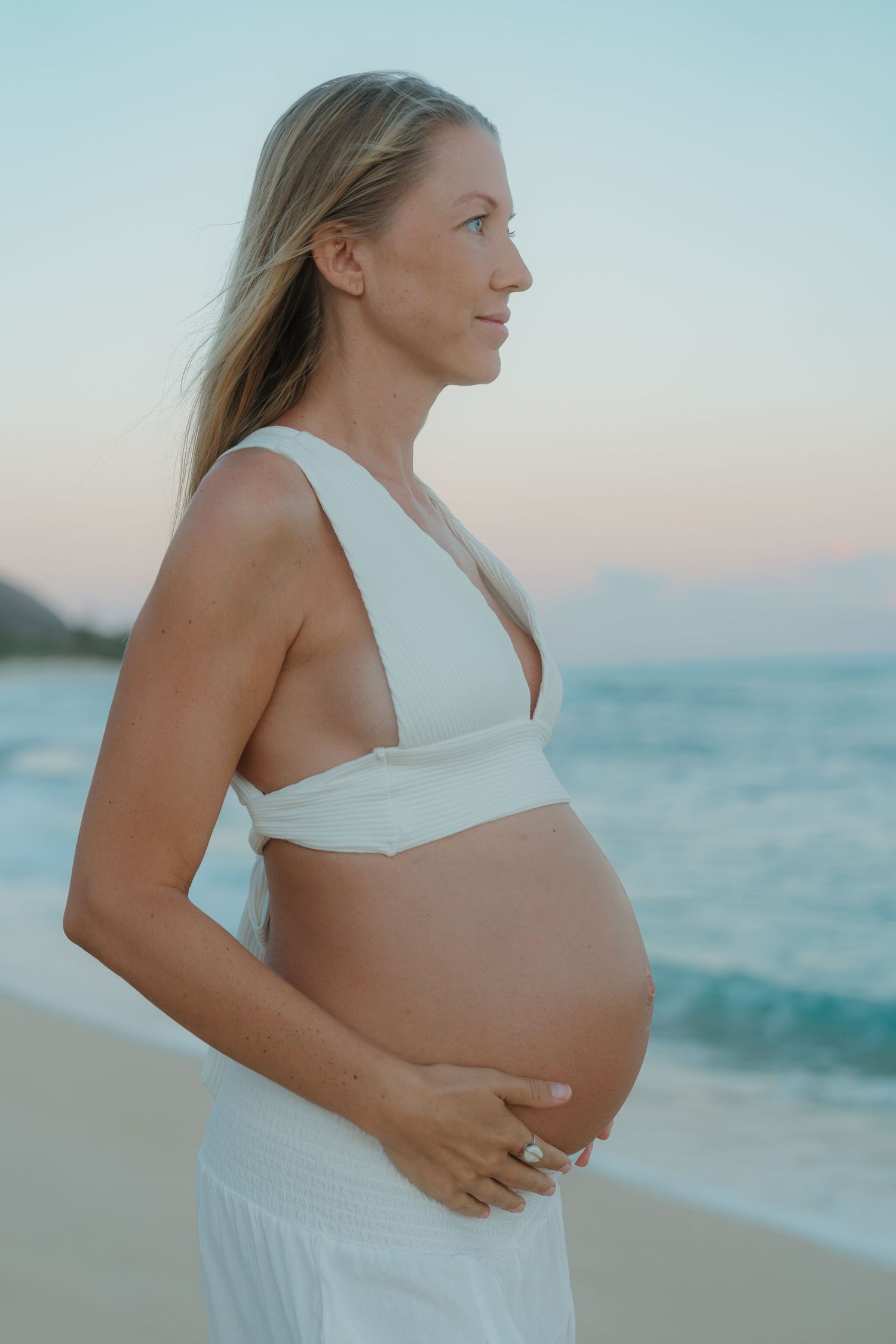 Pregnant woman in white bikini top cradling baby bump on a serene beach at sunset.