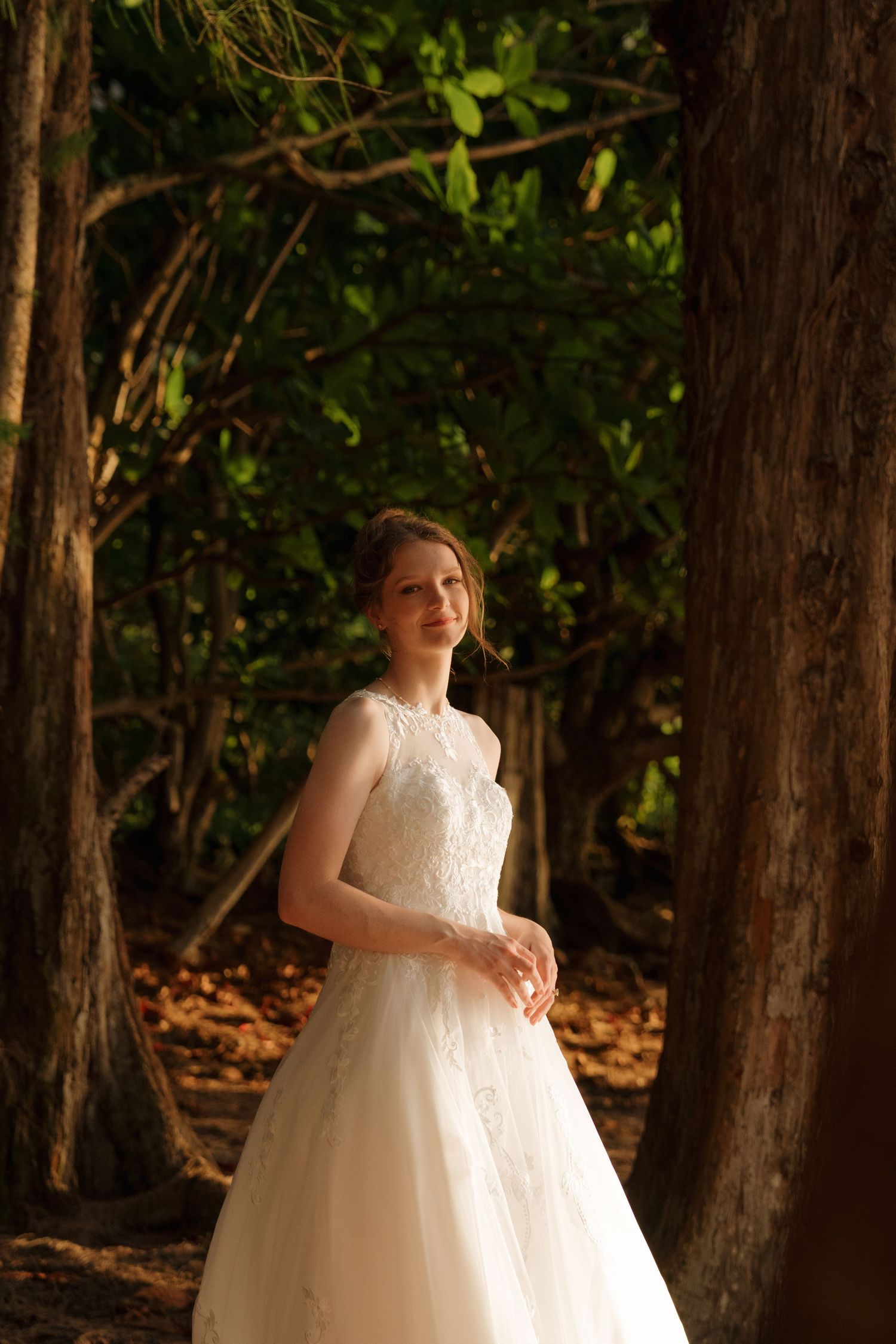 Bride in white lace wedding gown standing among tall trees in a sunlit forest setting.