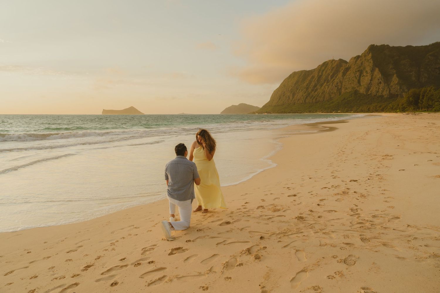 Man proposing on one knee to woman in yellow dress on sandy Hawaiian beach at golden sunset.