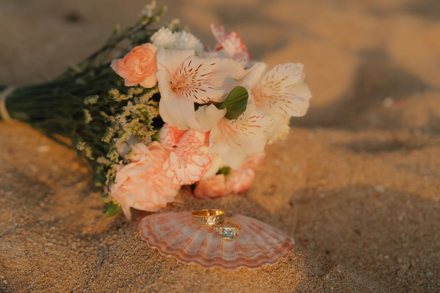 Bridal bouquet of peach flowers with wedding rings on a seashell resting on sandy beach at golden hour.