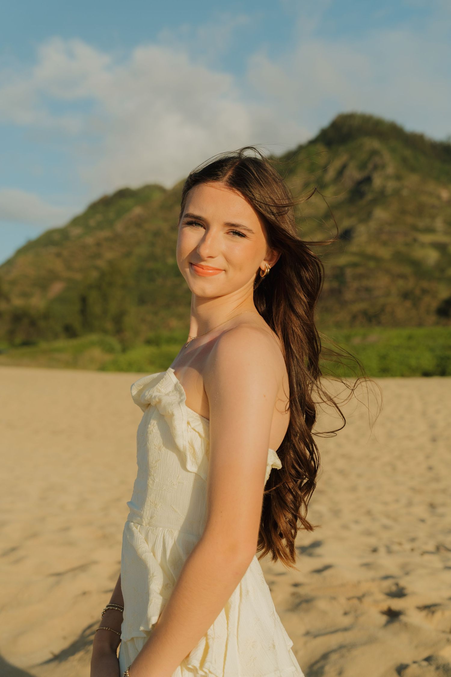Young woman in white dress smiling on sandy beach with green mountain and blue sky backdrop.