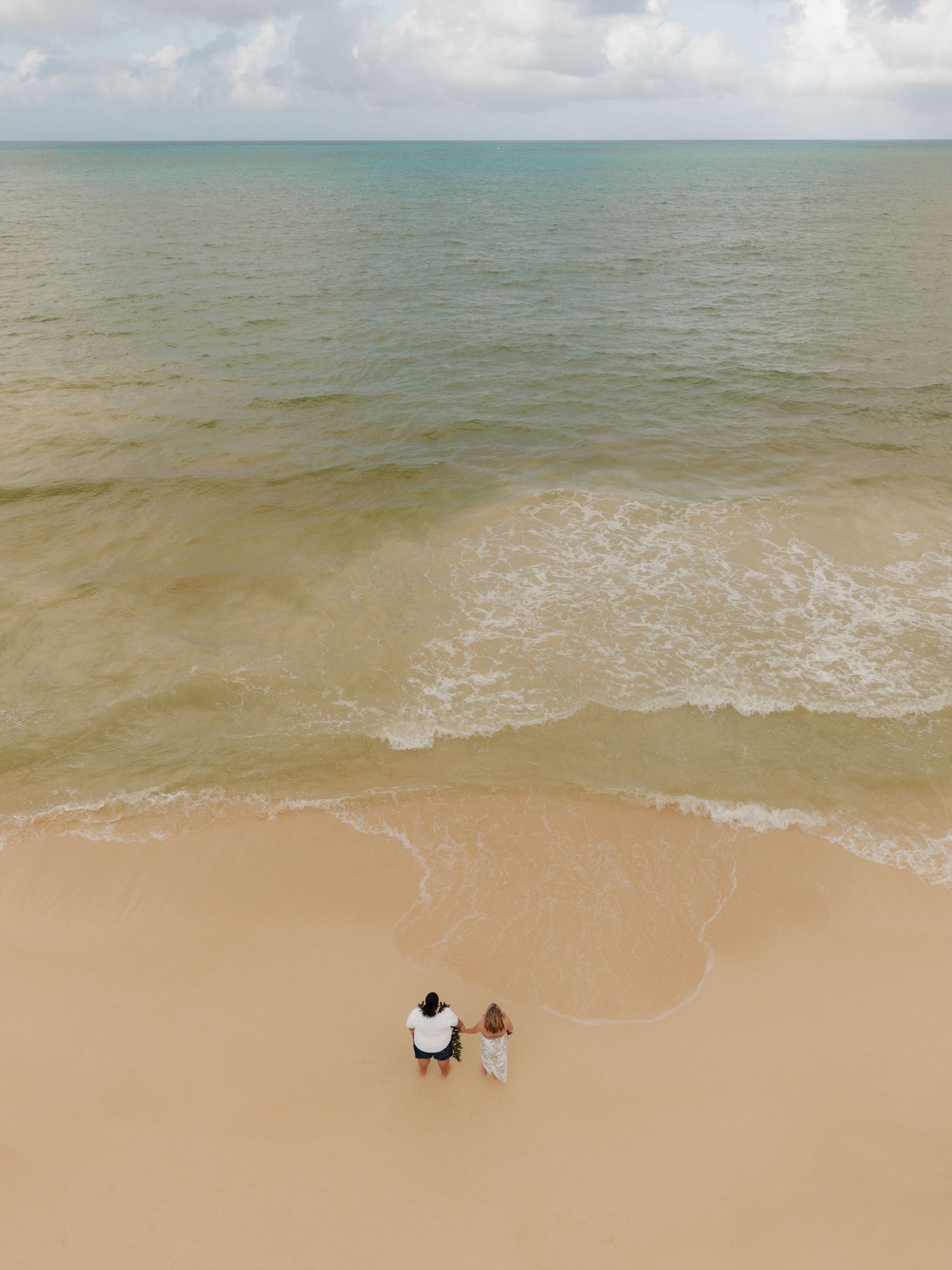Aerial view of a couple sitting together on a sandy beach shore with gentle ocean waves washing around them.