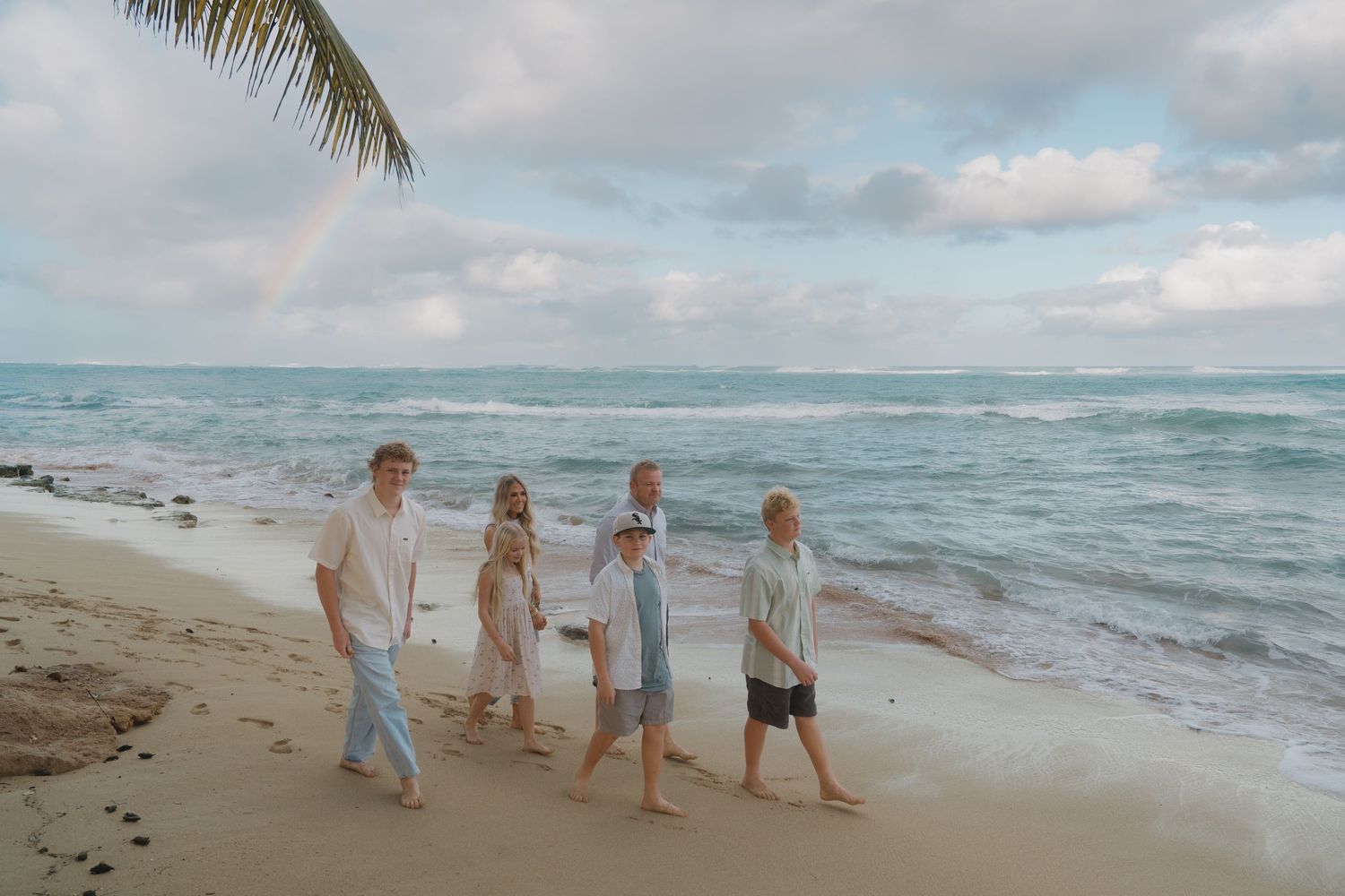 Family of six walking along sandy beach with rainbow visible over the ocean horizon.