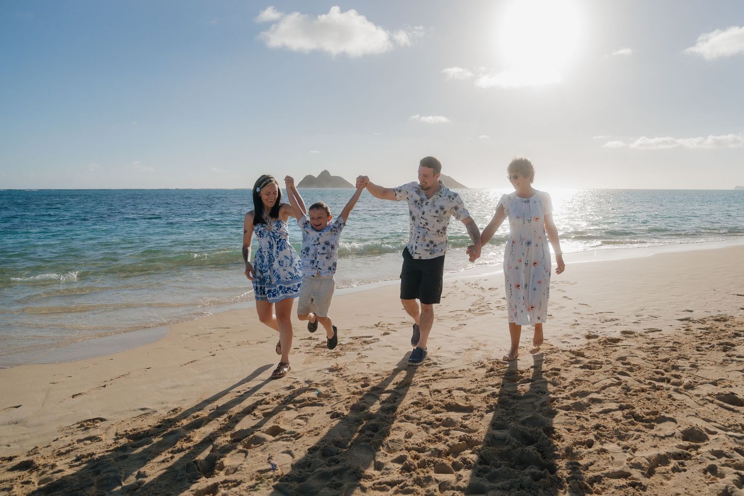 Joyful family of four holding hands and walking along a sunny Hawaiian beach with ocean waves.