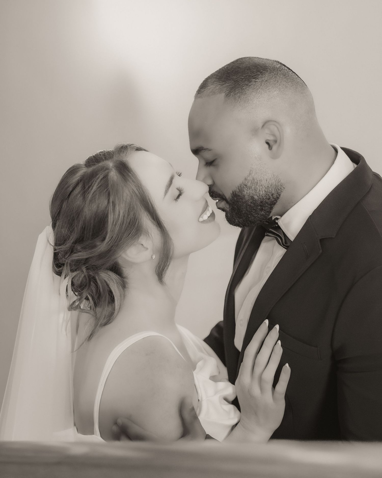 Black and white wedding photo of bride and groom sharing a romantic moment, nearly kissing, dressed in bridal gown and tuxedo.