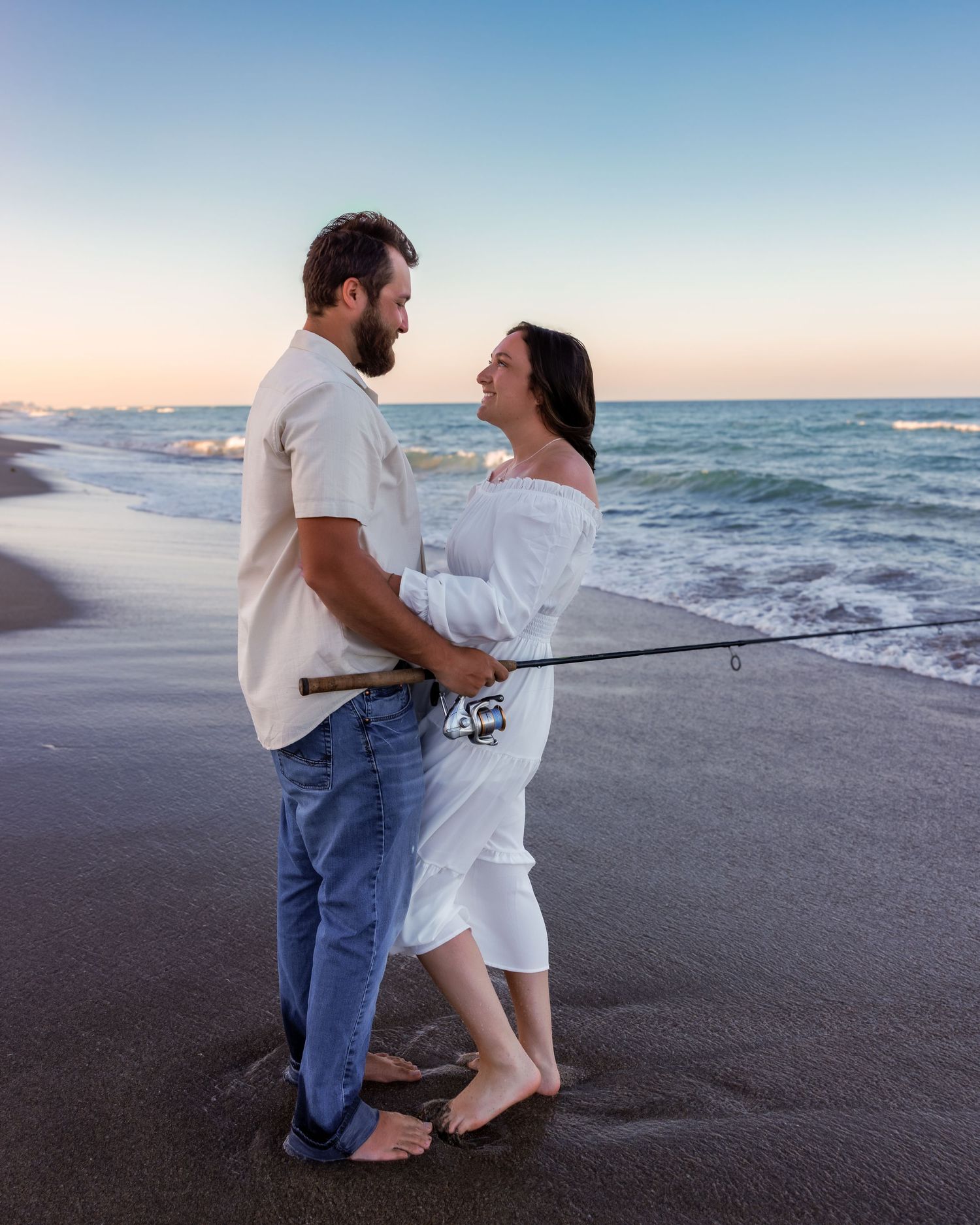Couple embracing on a beach at sunset, holding a fishing rod, with ocean waves in the background.