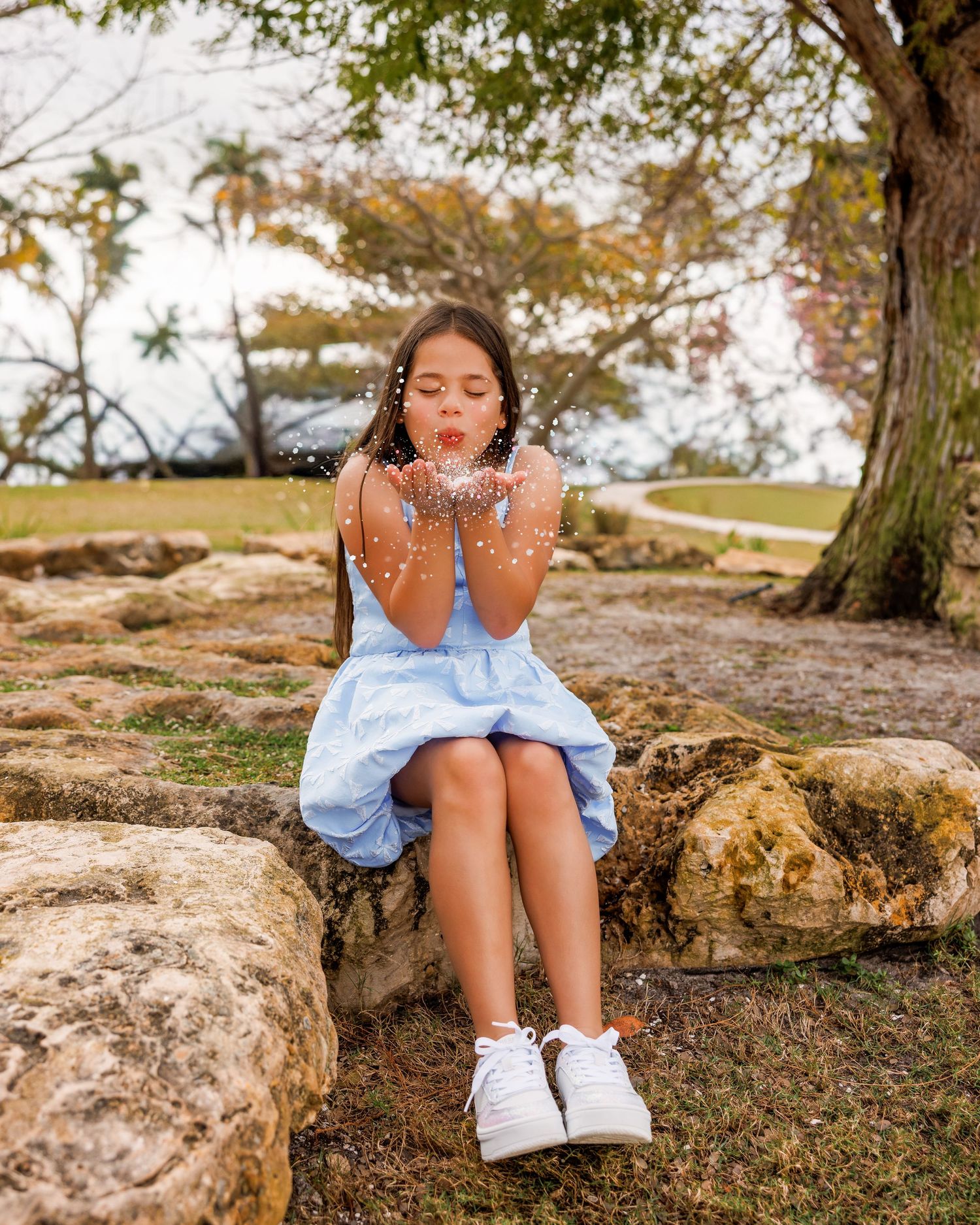 A young girl in a blue dress sits on rocks, blowing glitter from her hands in a lush outdoor park setting.