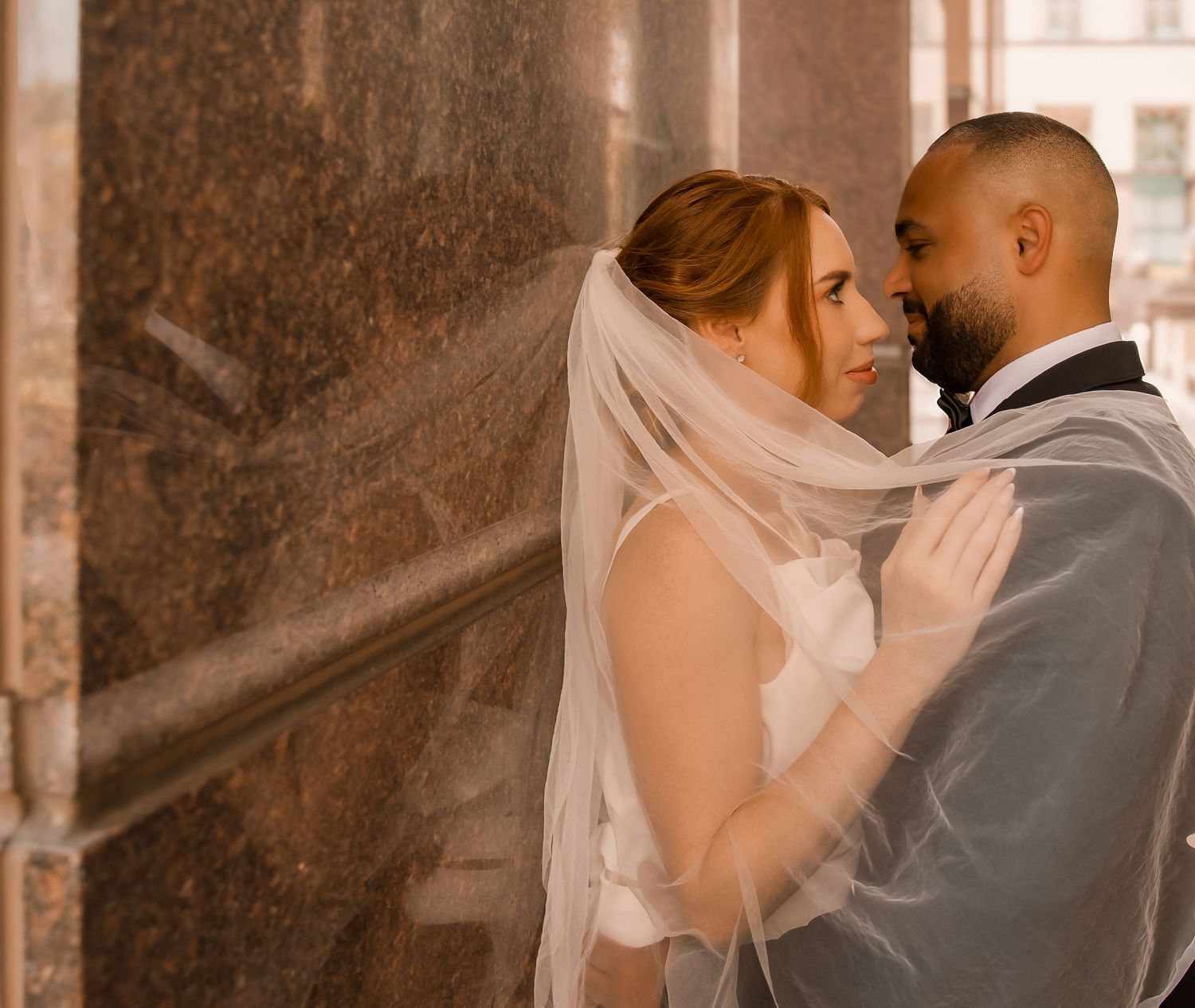 A bride and groom share a romantic moment near a marble wall, the bride wearing a flowing veil on their wedding day.