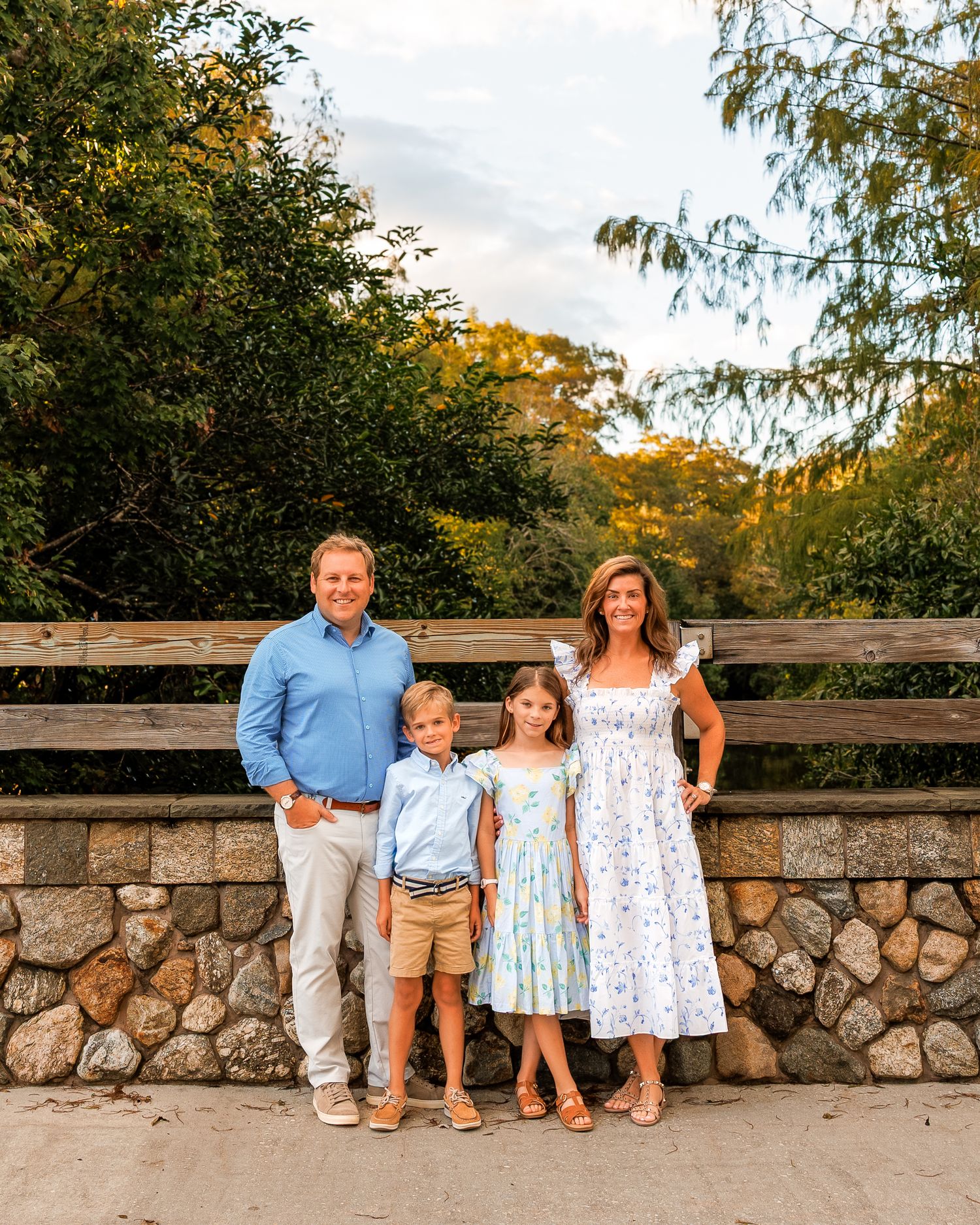 Family of four posing outdoors near a stone wall surrounded by lush green trees during golden hour.