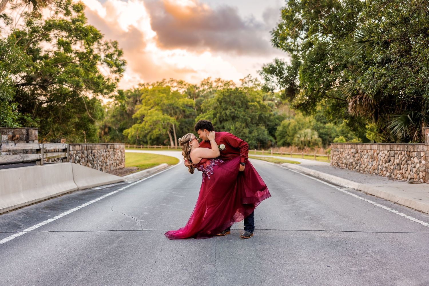 Couple in red formal wear sharing a romantic dip kiss on a road at sunset with lush trees in background.