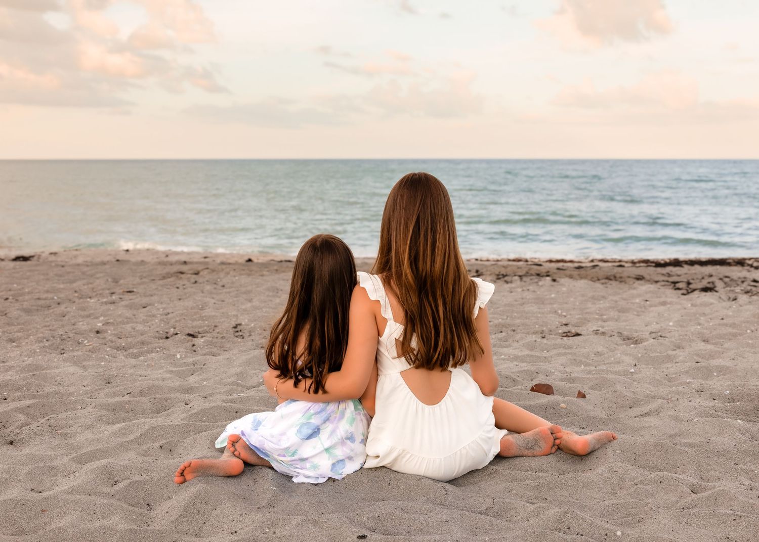 Two girls with long brown hair sit together on a sandy beach facing the calm ocean at sunset.