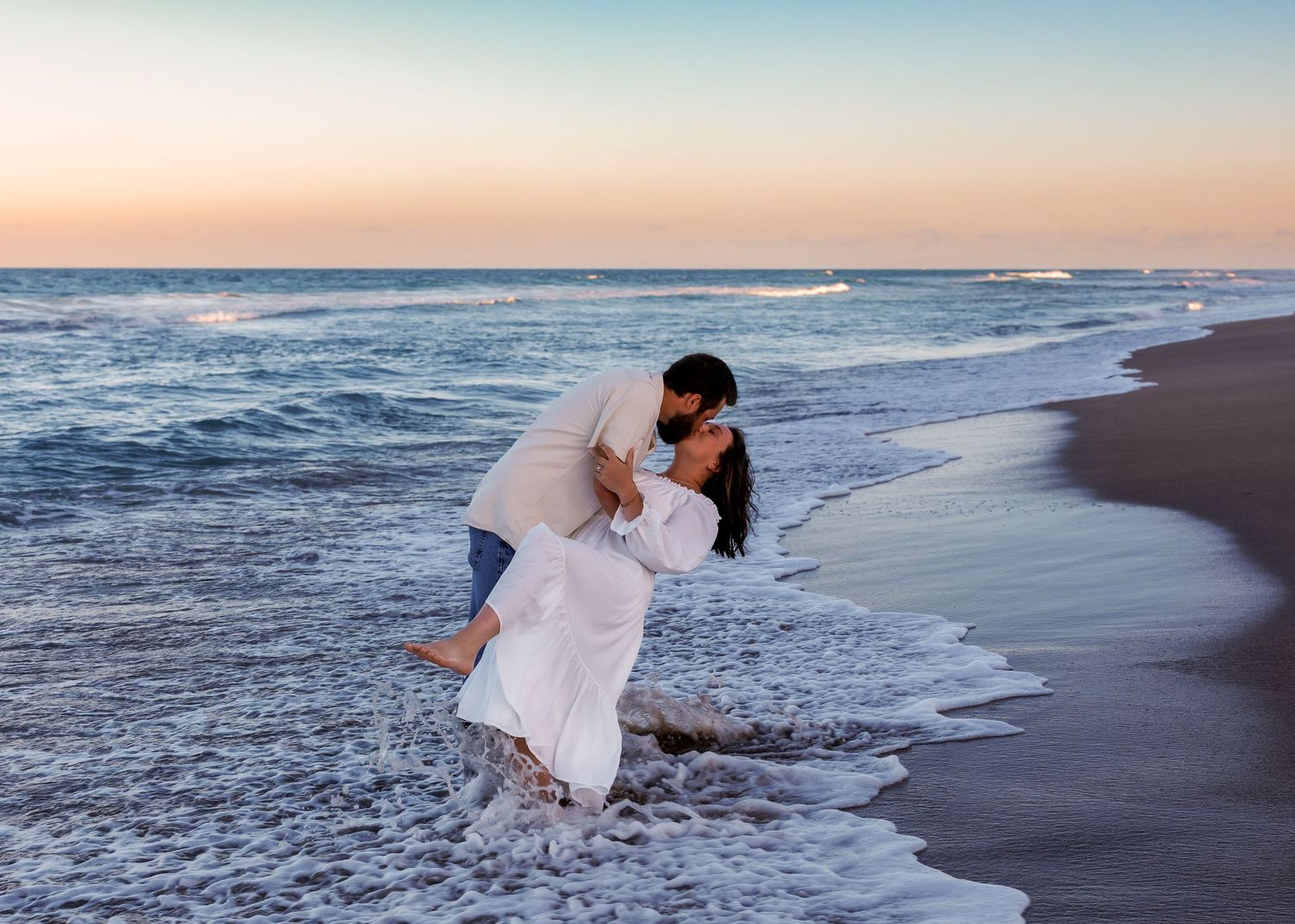 A romantic couple in white attire embraces on a beach at sunset as waves crash on the shore.
