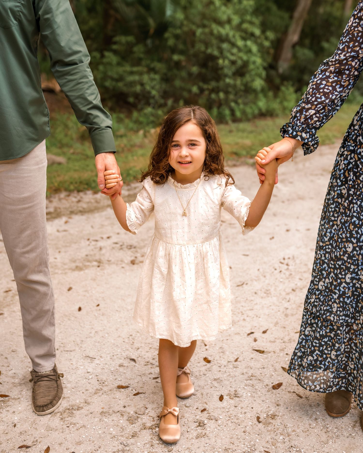 A family walking together on a dirt path holds hands in a line while wearing casual outdoor clothing.
