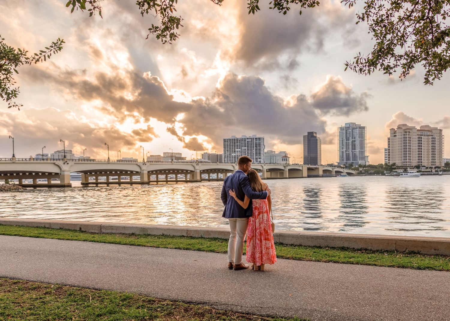 Couple standing on waterfront path at sunset with city skyline and bridge reflecting on calm water.