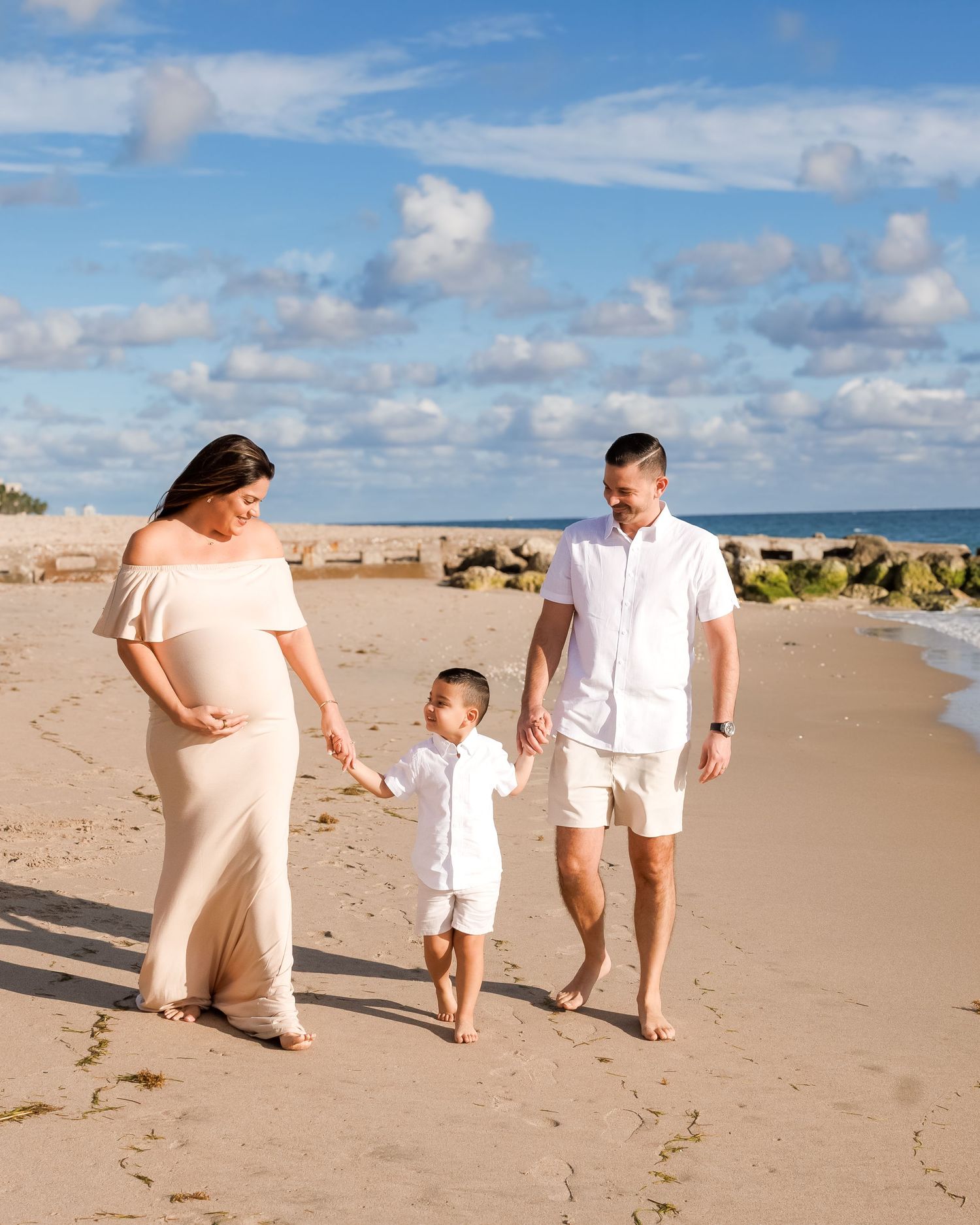 Pregnant woman in beige gown walks on beach with husband and toddler son under blue sky.